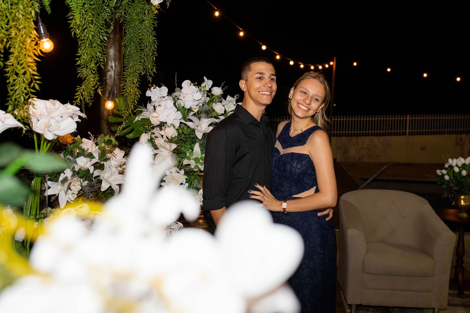 Couple posing together at an outdoor evening event with white floral decorations and string lights in the background.