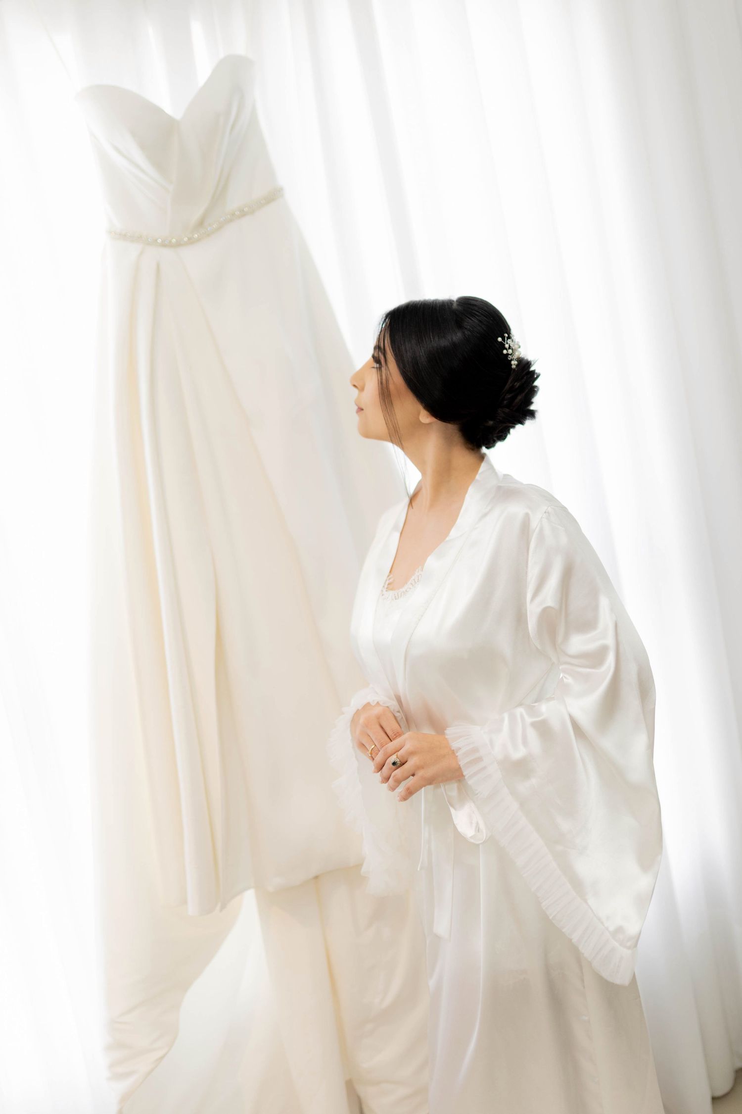 Bride in white satin robe admiring her elegant strapless wedding gown hanging by sheer curtains.