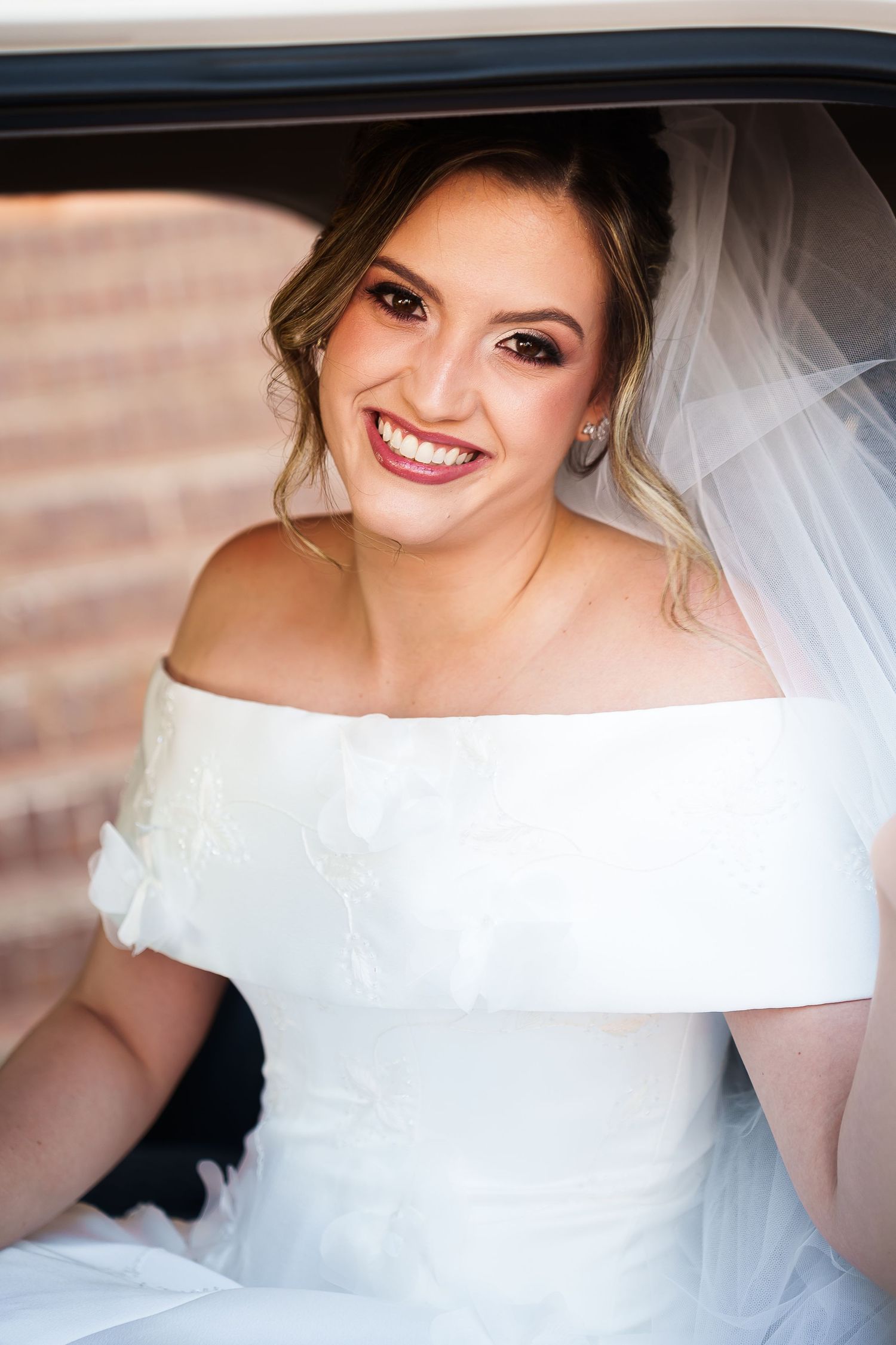 Smiling bride in white off-shoulder gown and veil sitting in a car window on her wedding day.