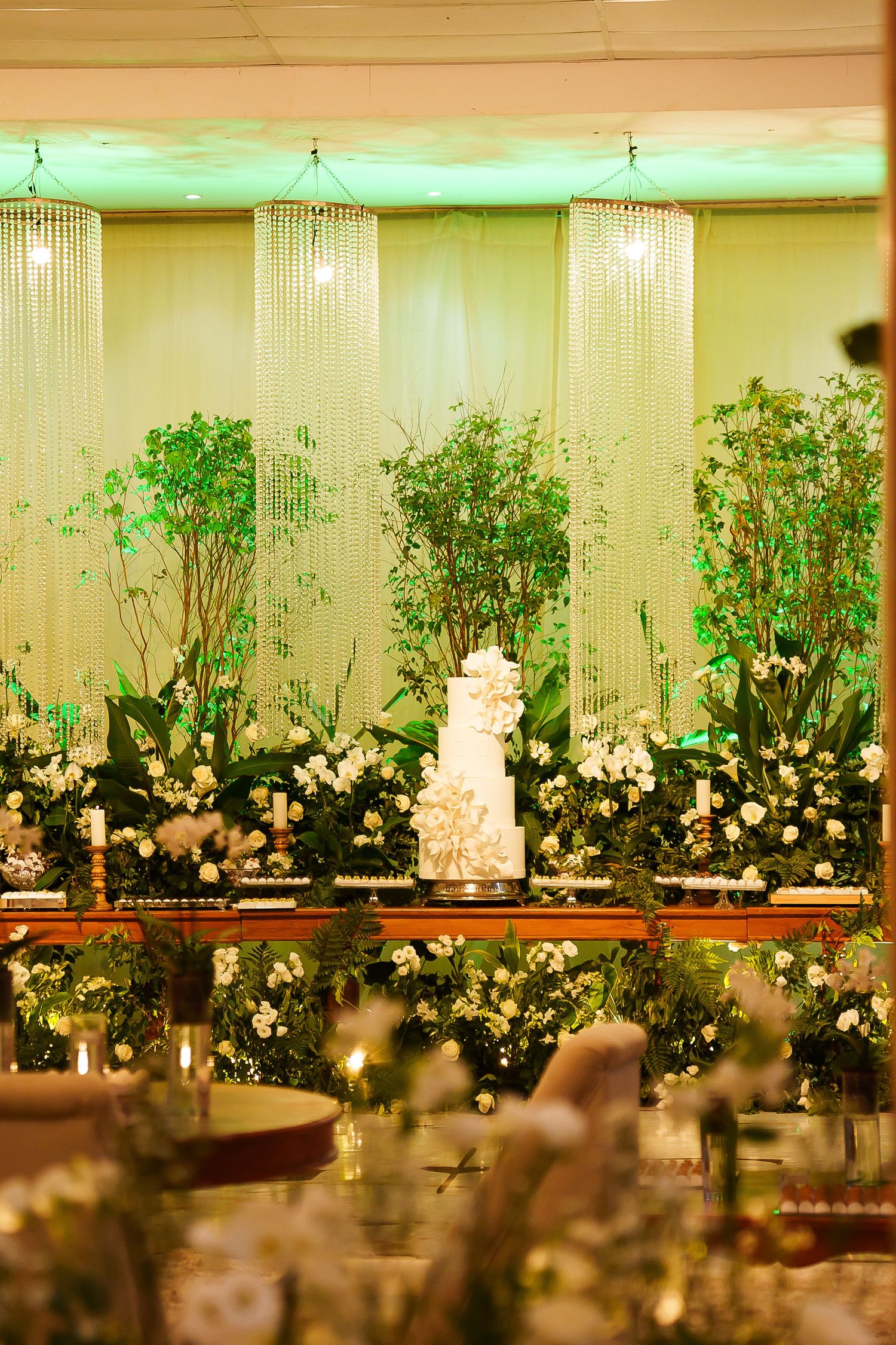 Elegant wedding reception with tiered white cake surrounded by lush greenery, white flowers, and crystal chandeliers in warm lighting.