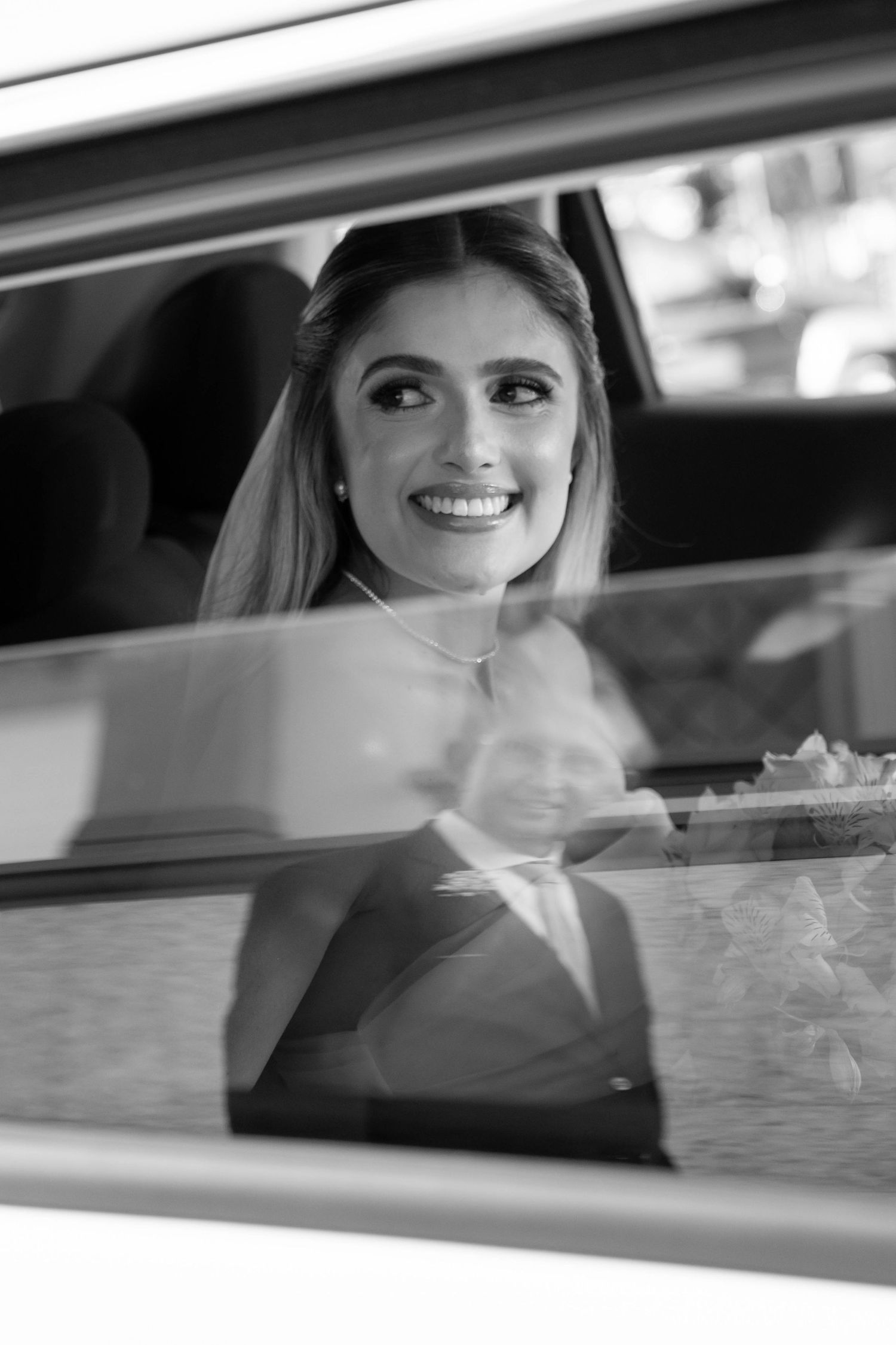 Black and white photo of a smiling bride in a car, with a groom's reflection visible in the window.