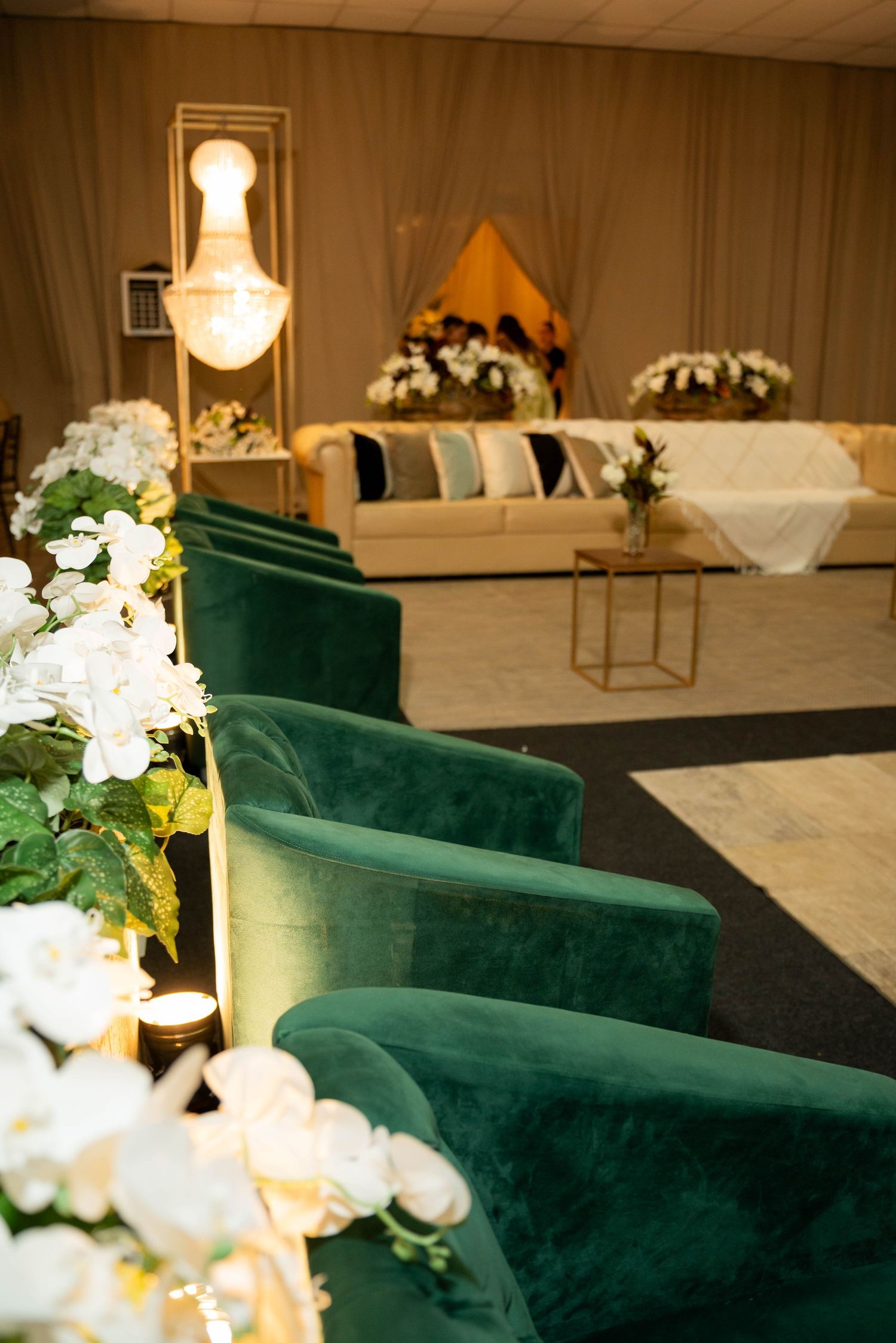 Elegant wedding venue with green velvet chairs lining an aisle, white floral arrangements, and a decorated sofa stage backdrop.