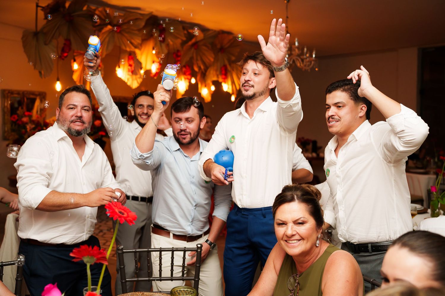 Men in white shirts celebrate at a festive party, raising bottles and cheering joyfully indoors.