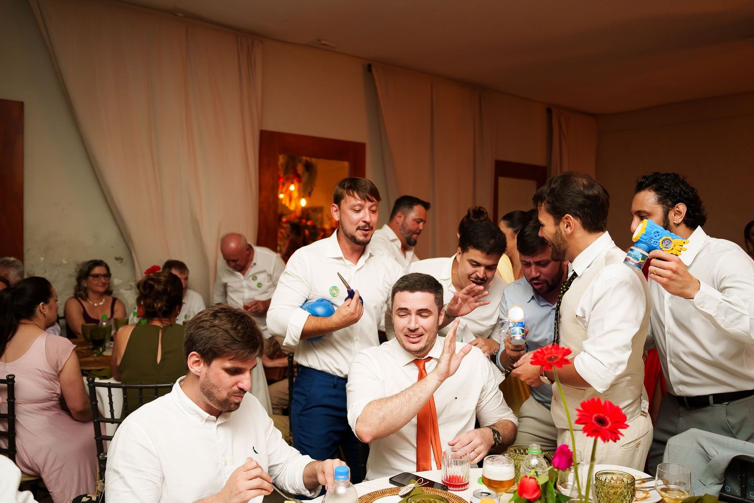 Group of men in dress shirts celebrating at a wedding reception dinner table, laughing and enjoying drinks.