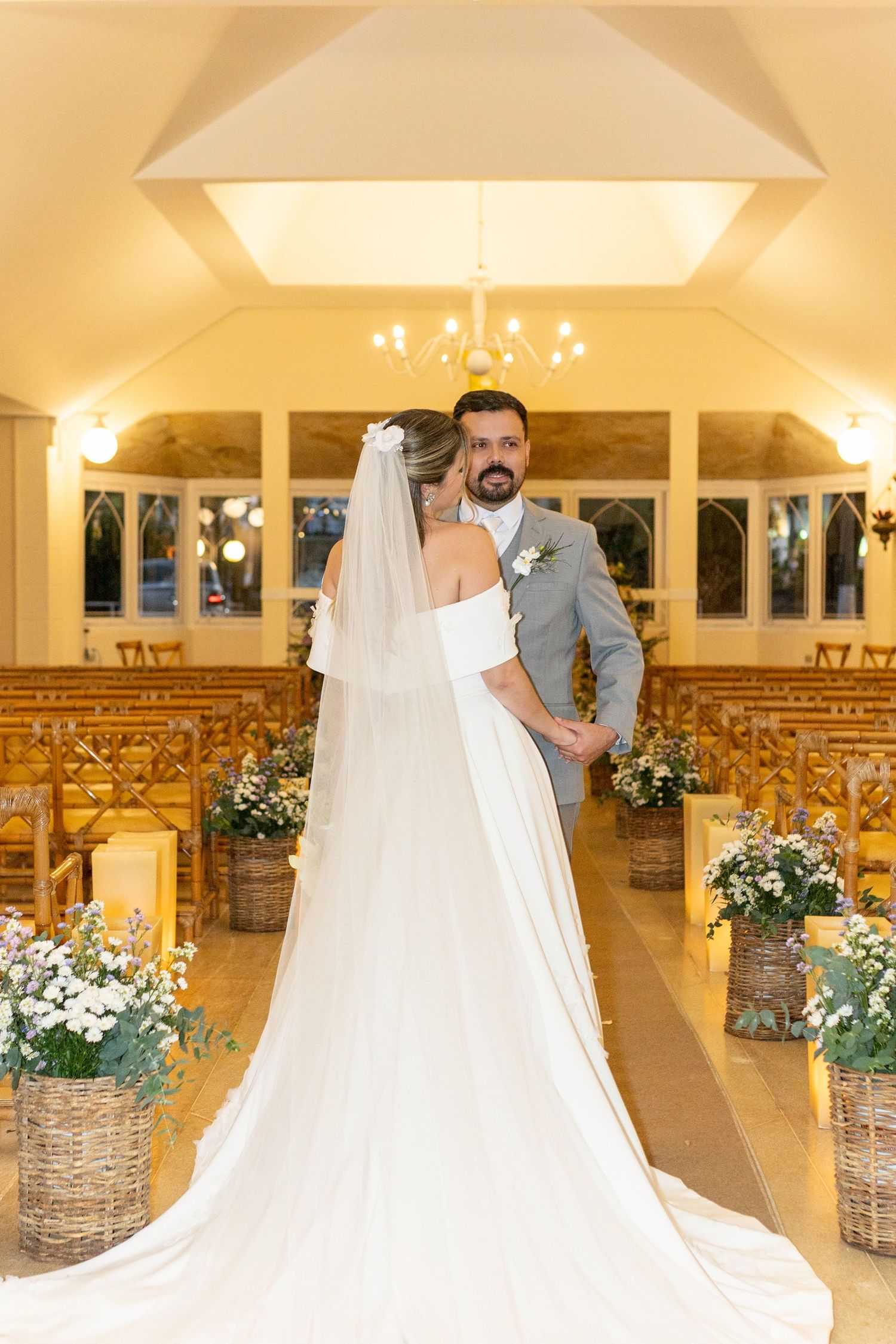 Bride and groom holding hands at the altar of an elegant chapel with floral arrangements and wooden chairs.