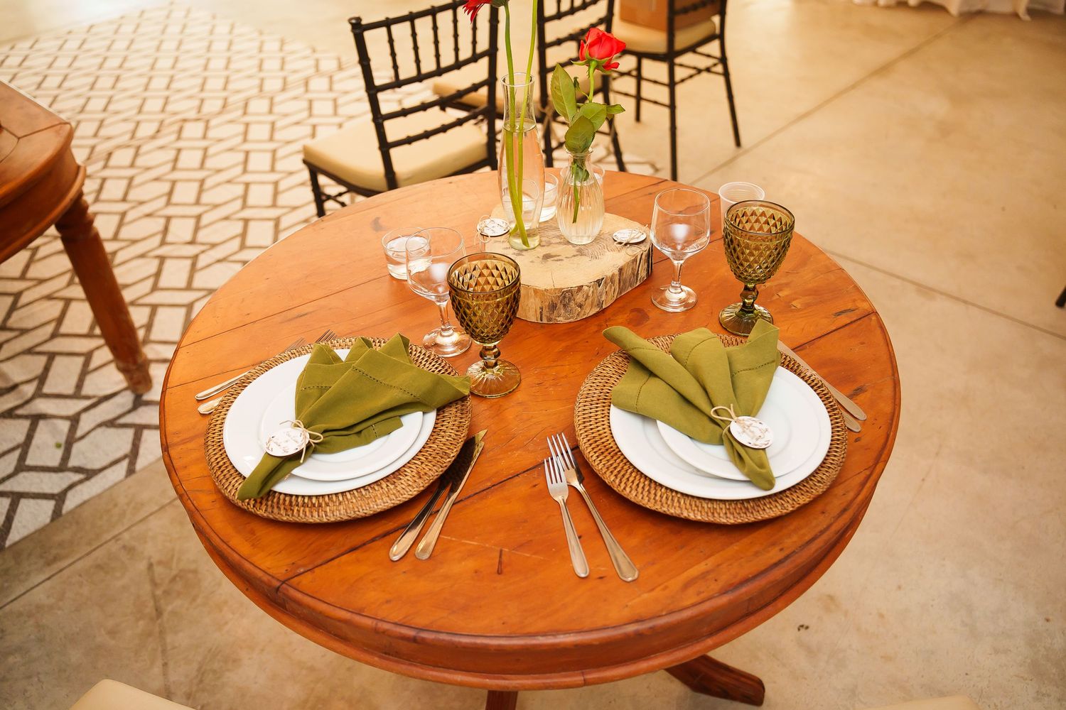 Round wooden table set for two with green napkins, gold goblets, white plates, and silverware for an elegant dining experience.