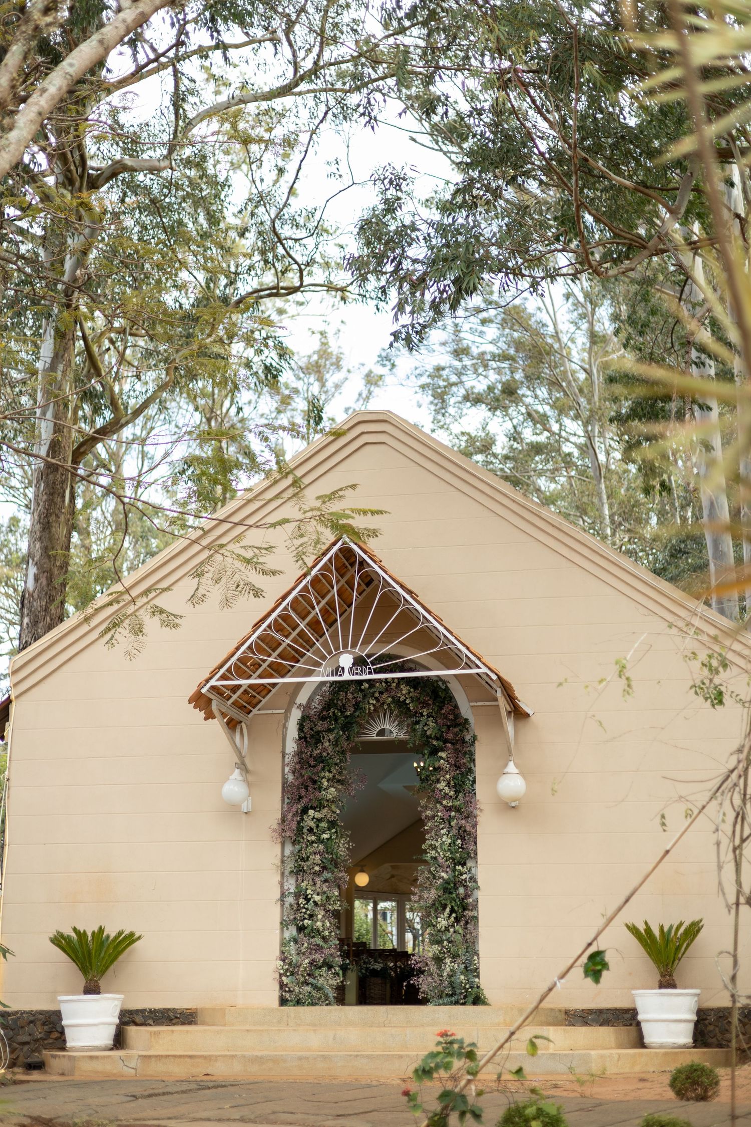 Charming cream chapel entrance adorned with lush greenery arch, globe lights, and decorative ironwork surrounded by tall trees.