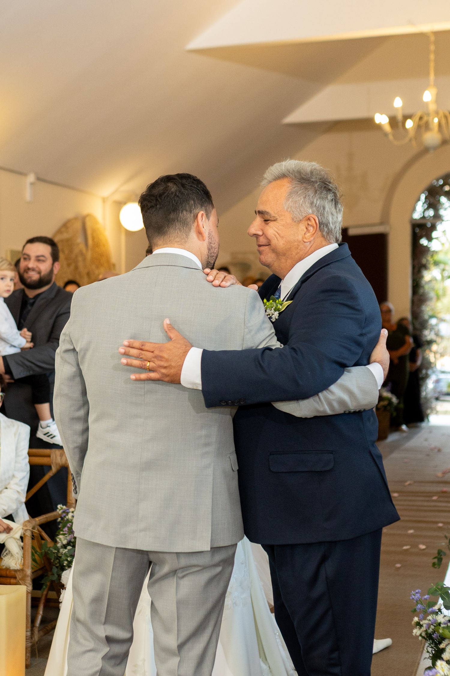 Two men in suits share a warm embrace during a wedding ceremony inside an elegant venue.