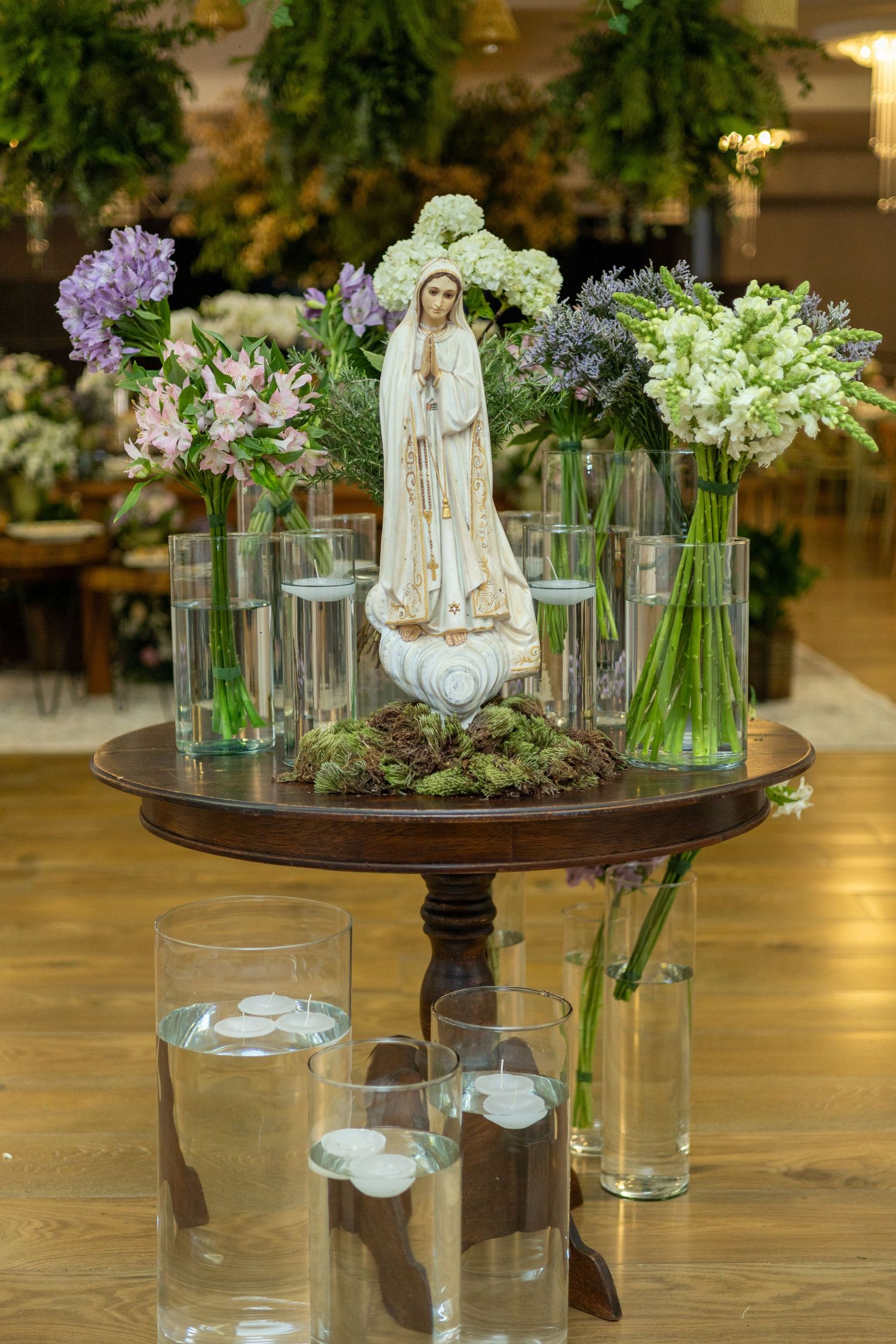 Our Lady of Fatima statue centerpiece surrounded by fresh flowers in glass vases on a wooden pedestal table.