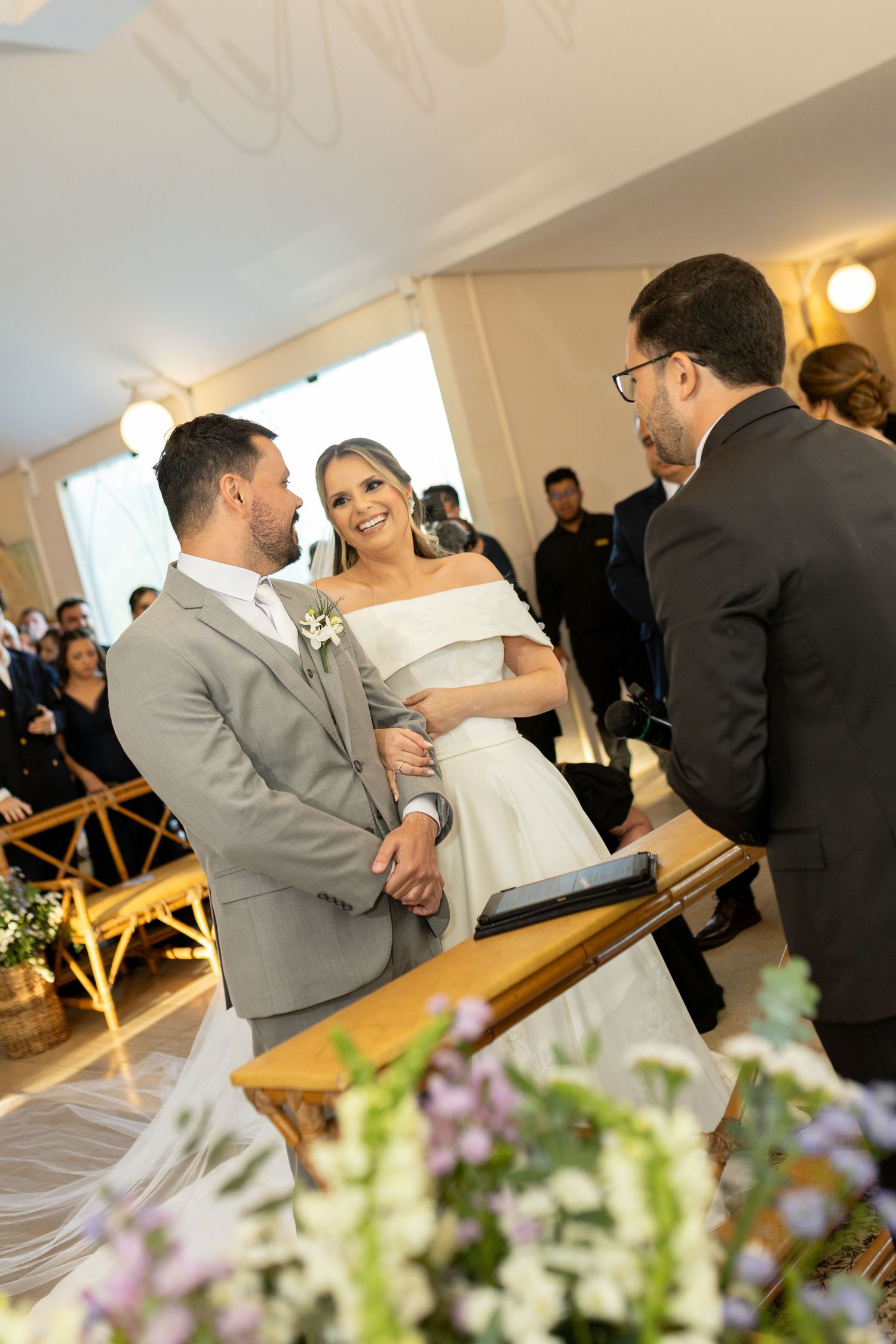 Bride and groom holding hands and smiling during wedding ceremony with officiant and guests in background.