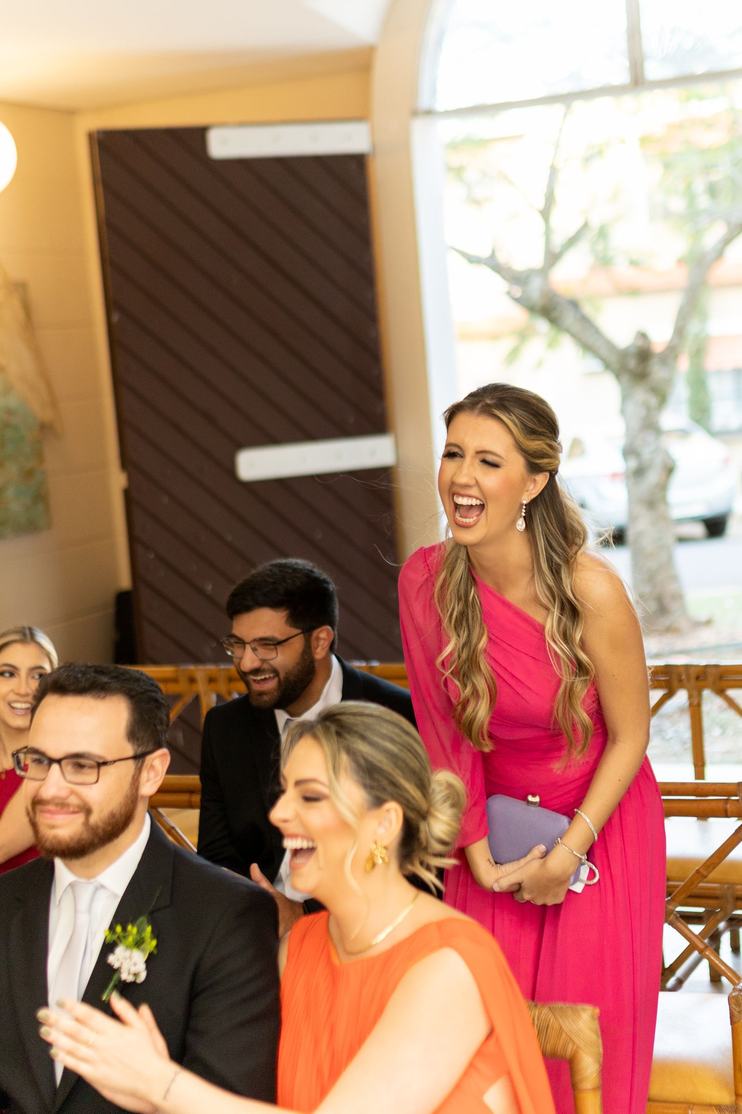 Laughing bridesmaid in pink gown stands at wedding ceremony with smiling guests in formal attire seated nearby.
