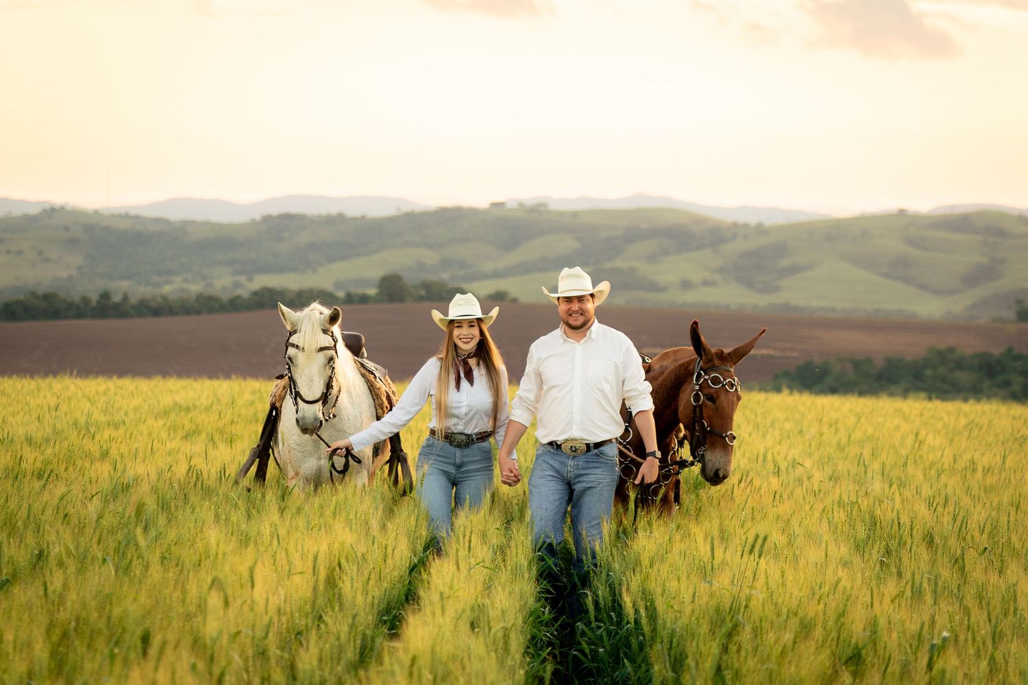 Couple in cowboy hats walking with horses through a green wheat field at sunset with rolling hills.