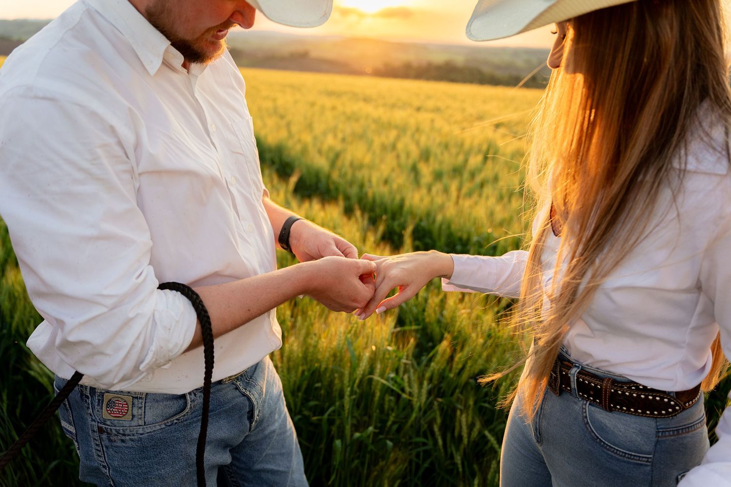 Couple in white shirts holding hands in a golden wheat field at sunset, wearing cowboy hats and jeans.