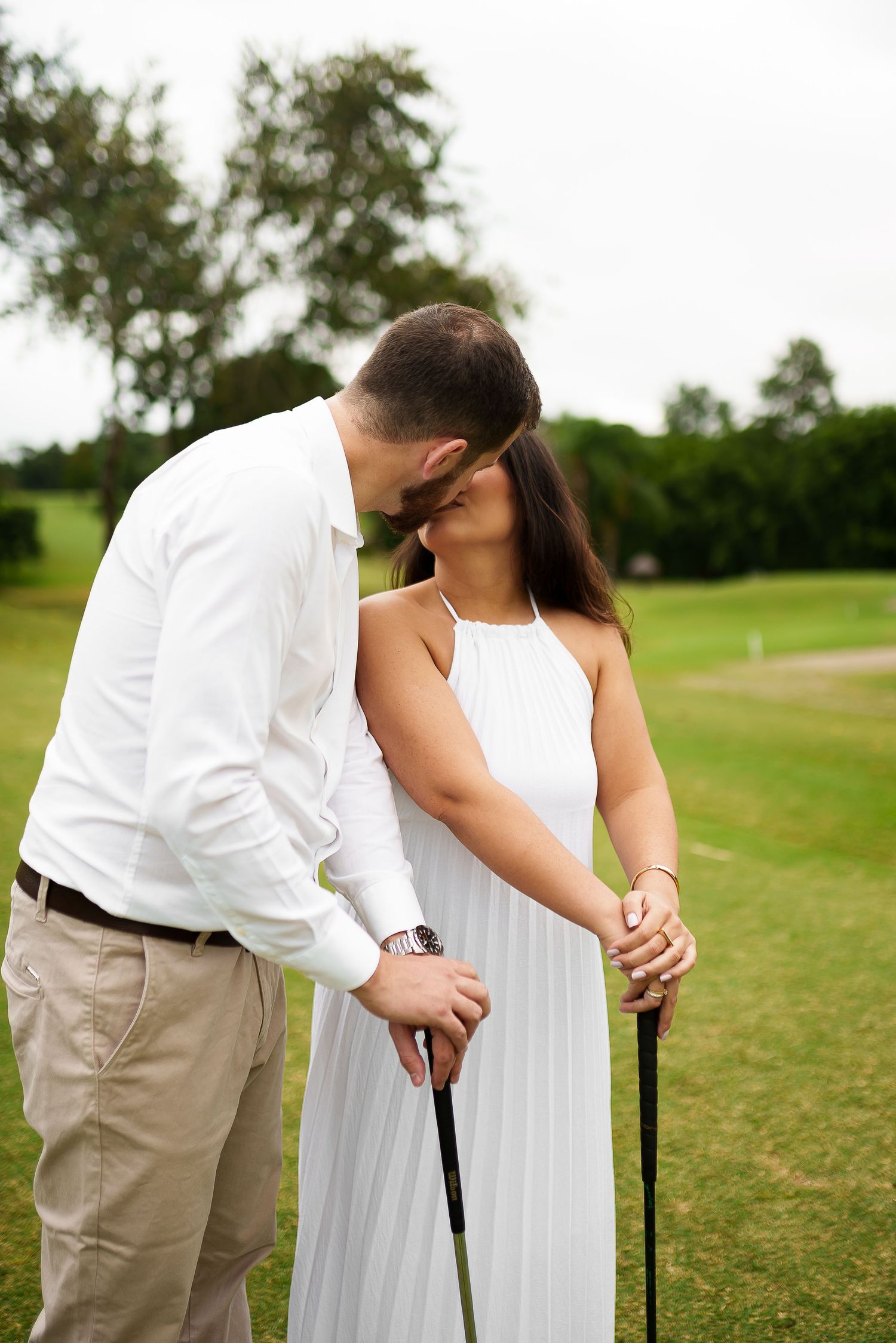 Couple sharing a romantic kiss on a golf course, woman in white dress holding a golf club on a lush green fairway.