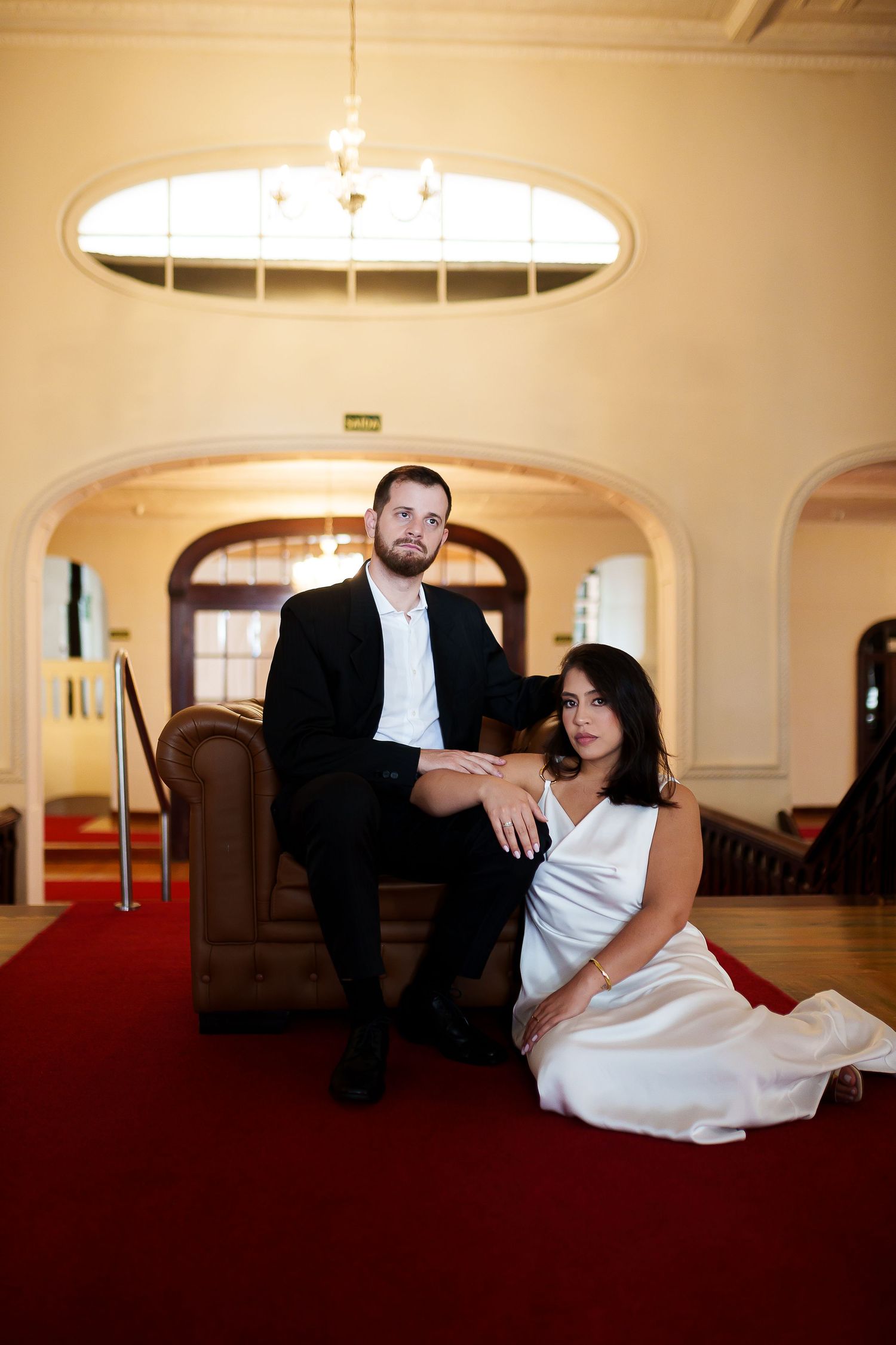 Elegant couple poses in grand hall, man in black suit sits in chair, woman in white dress sits on red carpet.
