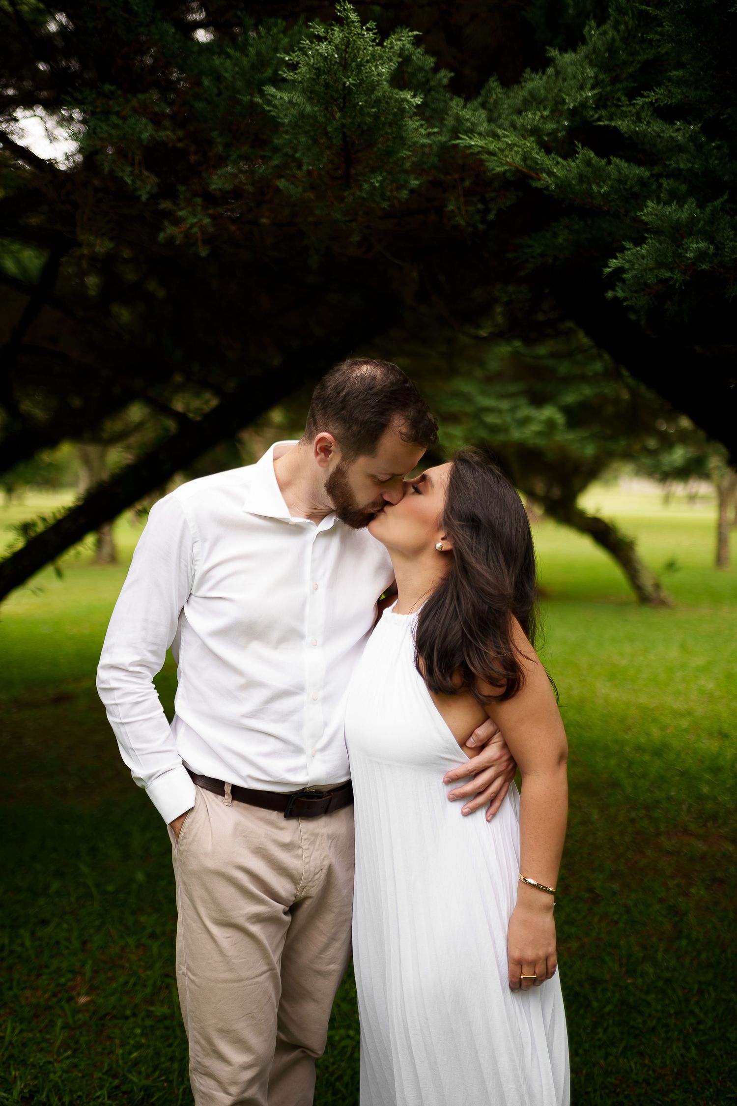 Couple sharing a romantic kiss outdoors in a lush green park, wearing white dress and white shirt.
