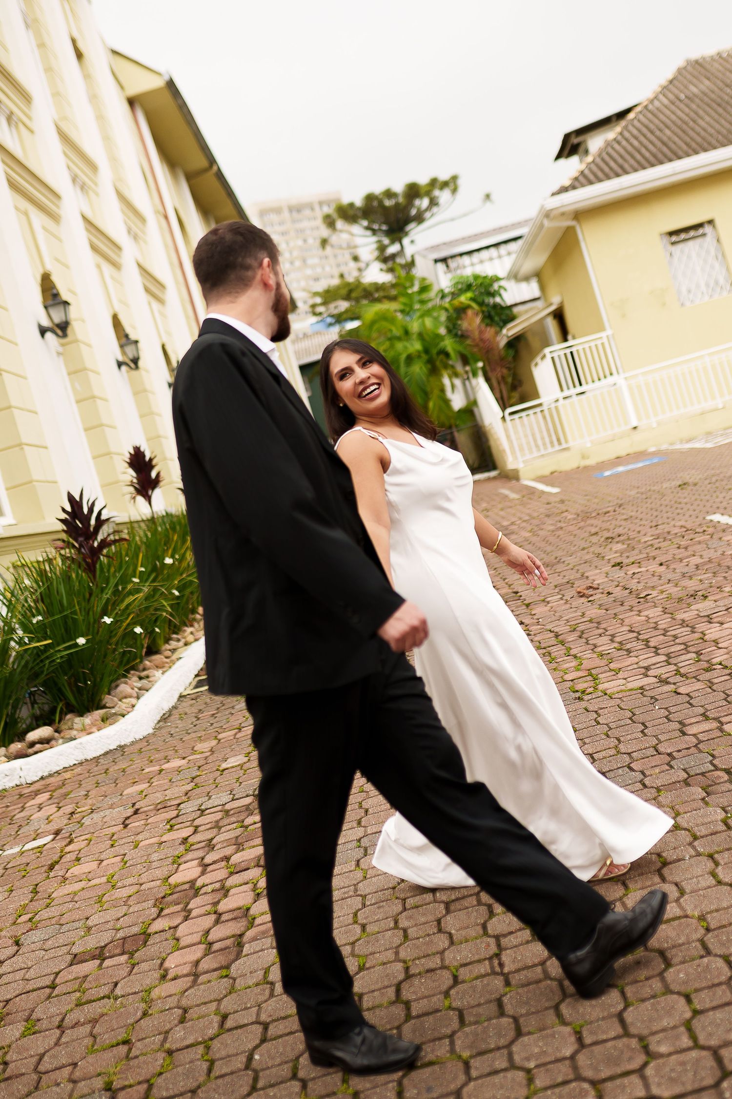 Happy bride in white dress laughing while walking hand in hand with groom in black suit on cobblestone path.