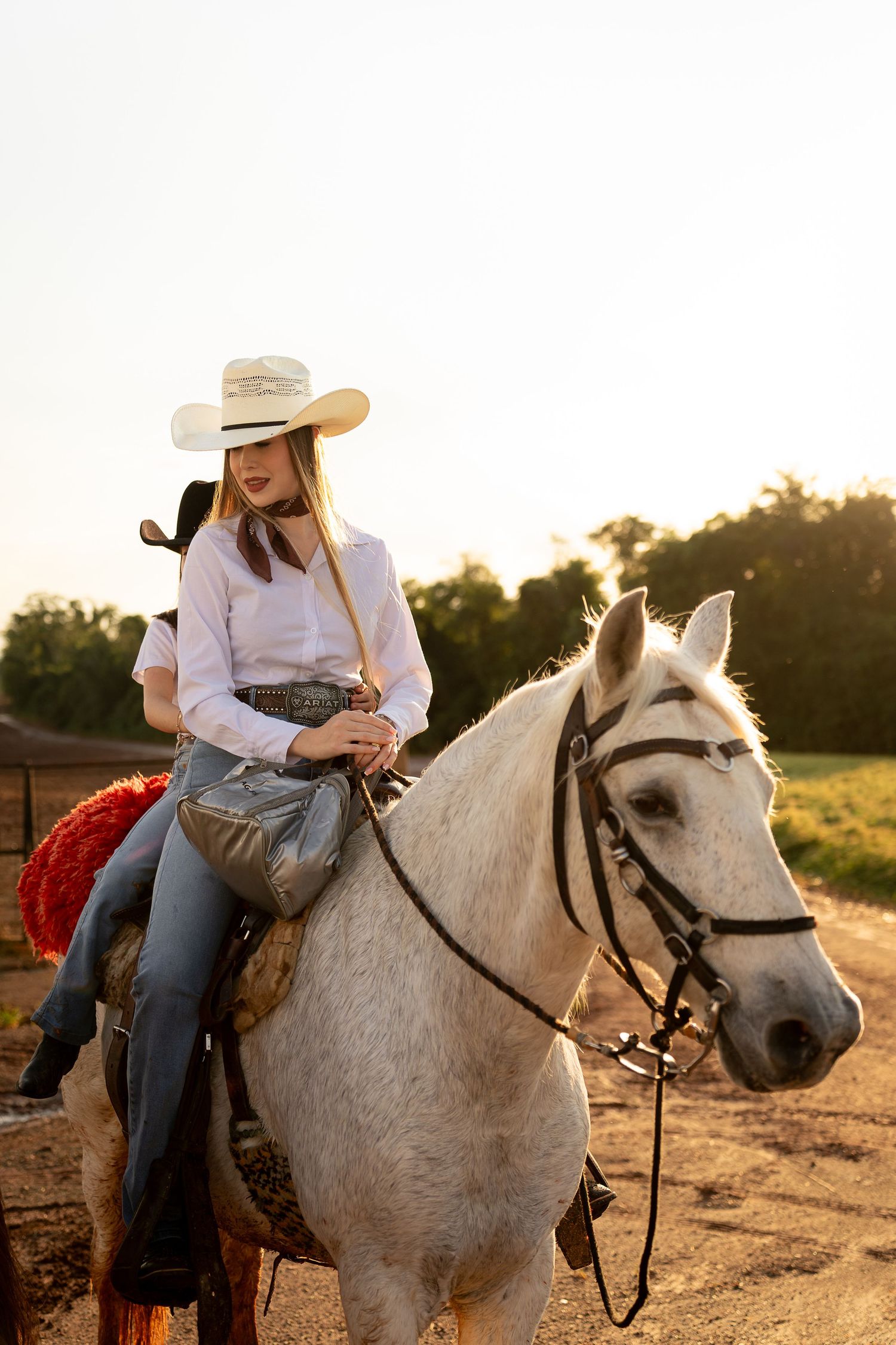 Young woman in white cowboy hat riding a white horse on a sunny farm road at golden hour.