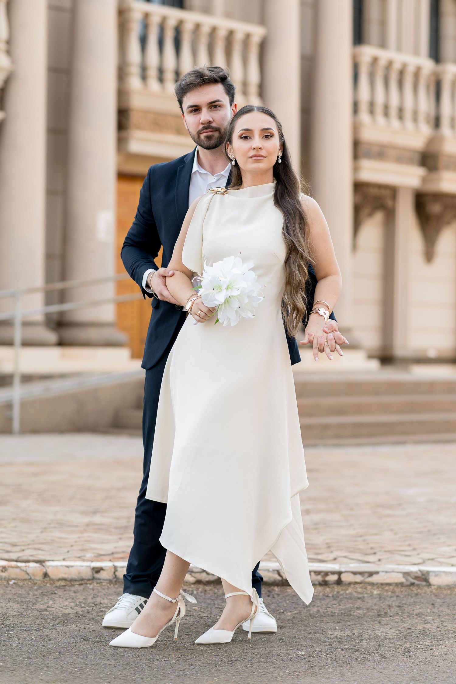 Bride in white midi dress holding bouquet poses with groom in navy suit outside historic building.