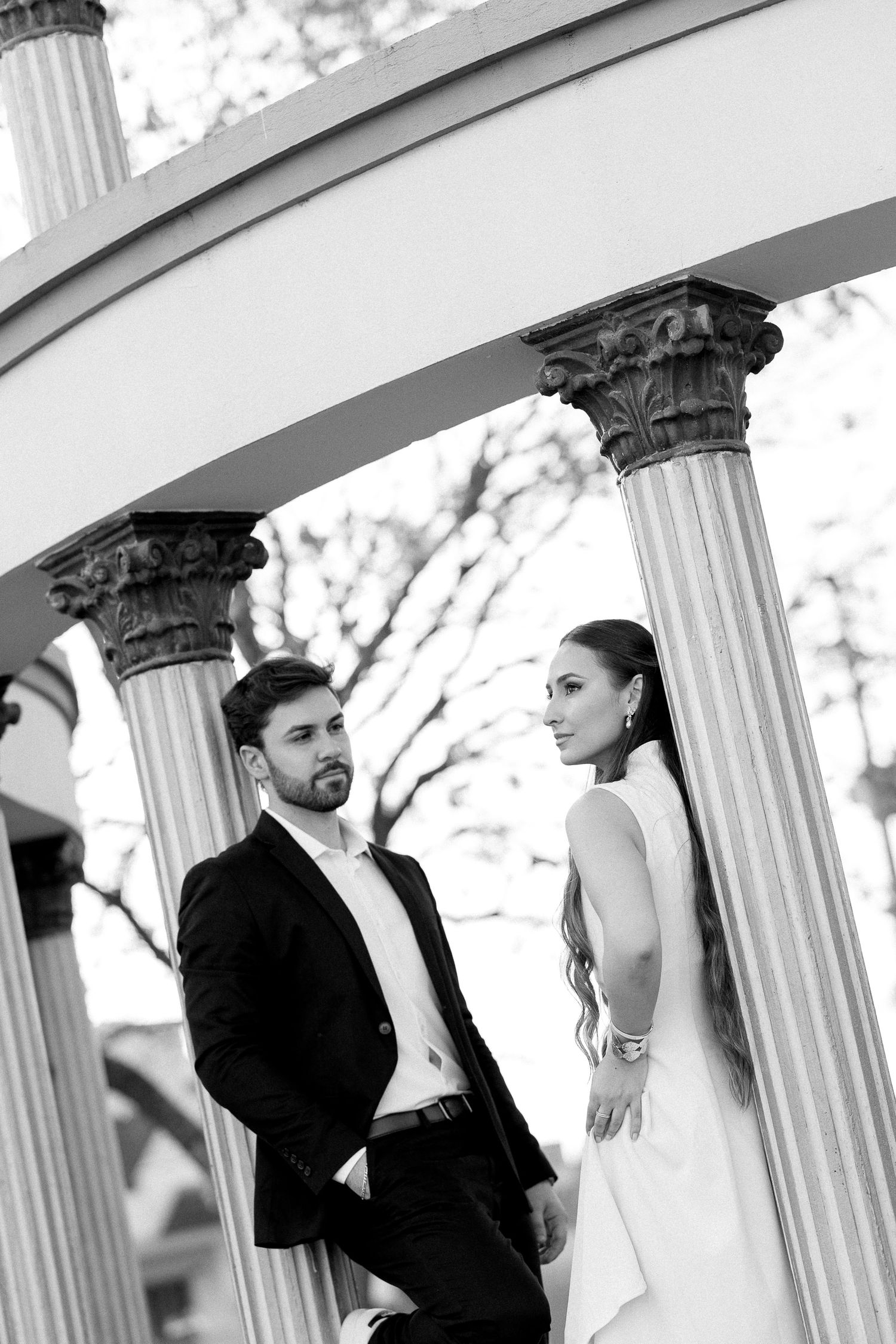 Black and white photo of elegant bride and groom posing between classical columns in a romantic outdoor setting.