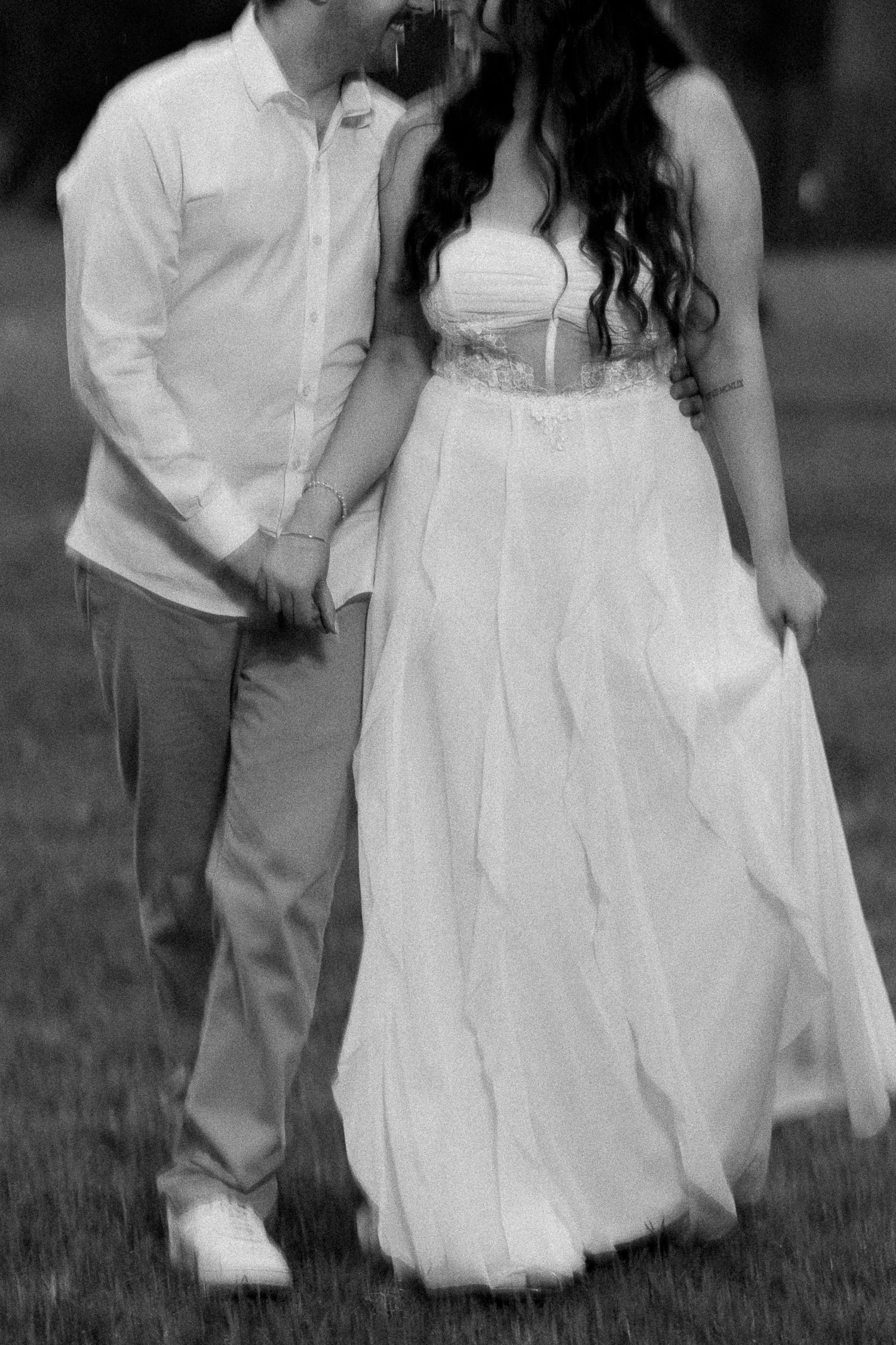 Black and white photo of a couple holding hands, woman in flowing white dress, man in white shirt walking outdoors.