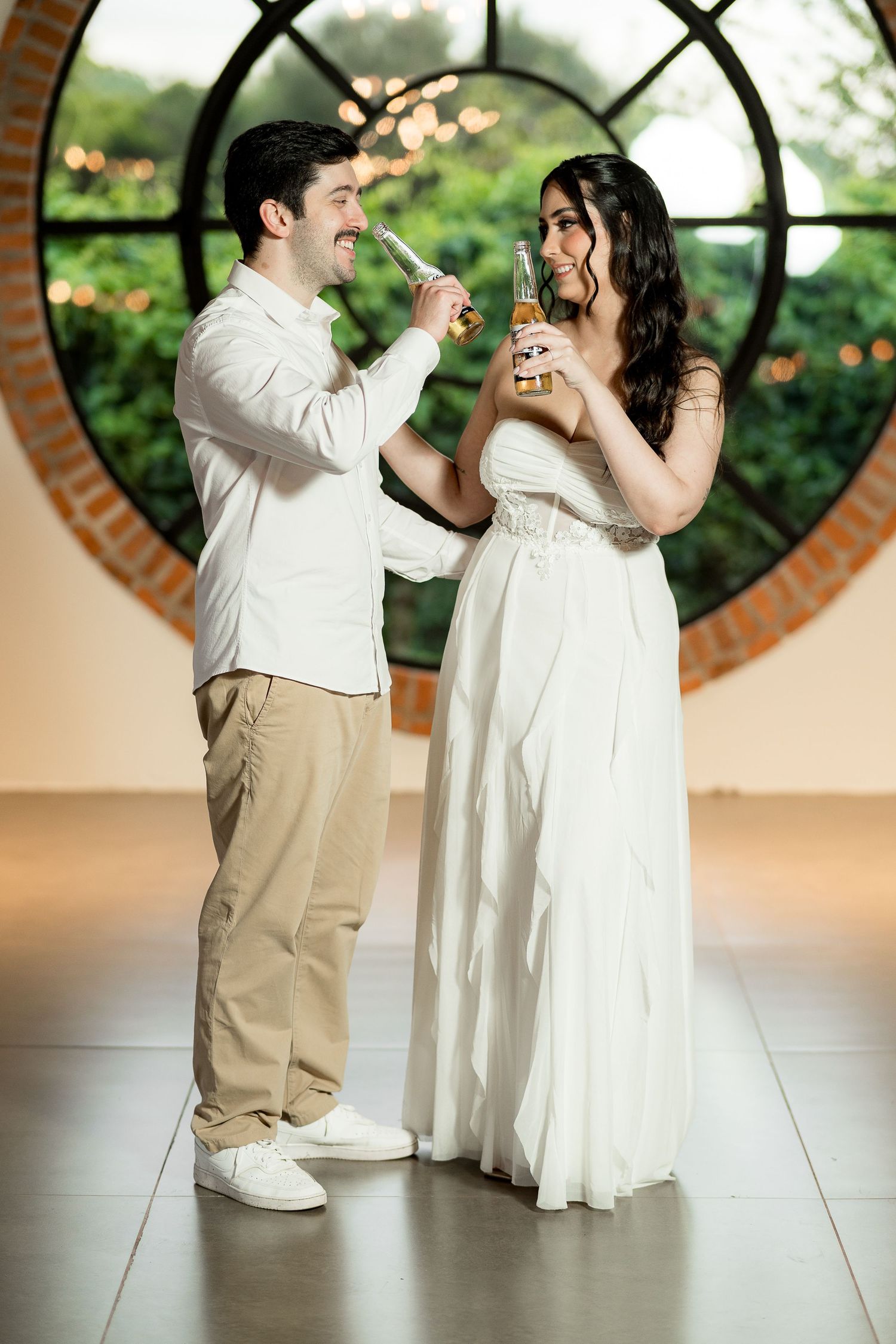 Bride in white gown and groom in white shirt clink beer bottles, smiling at each other in elegant venue.