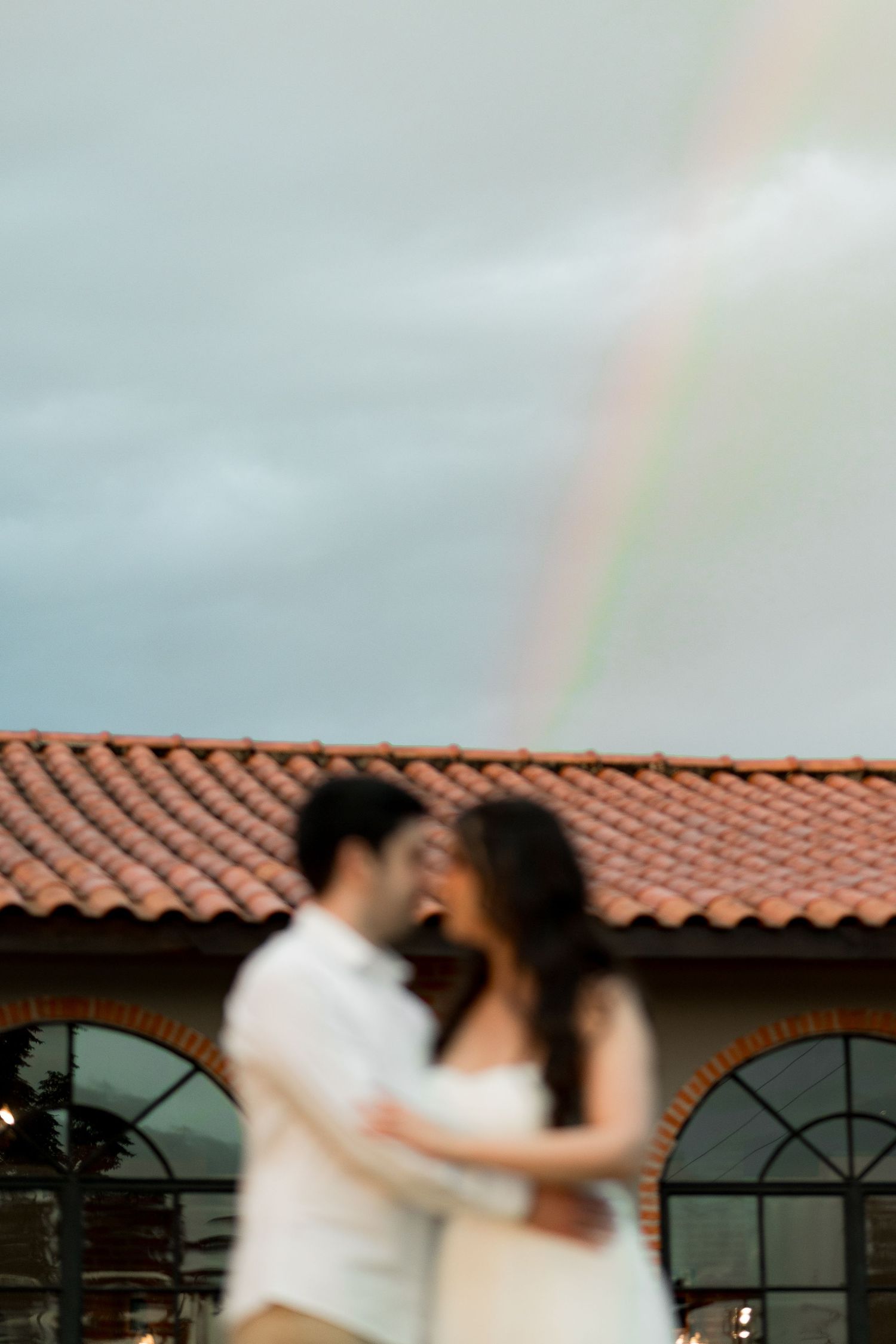 Blurred couple embracing romantically outdoors with a rainbow arching over a terracotta roof building.