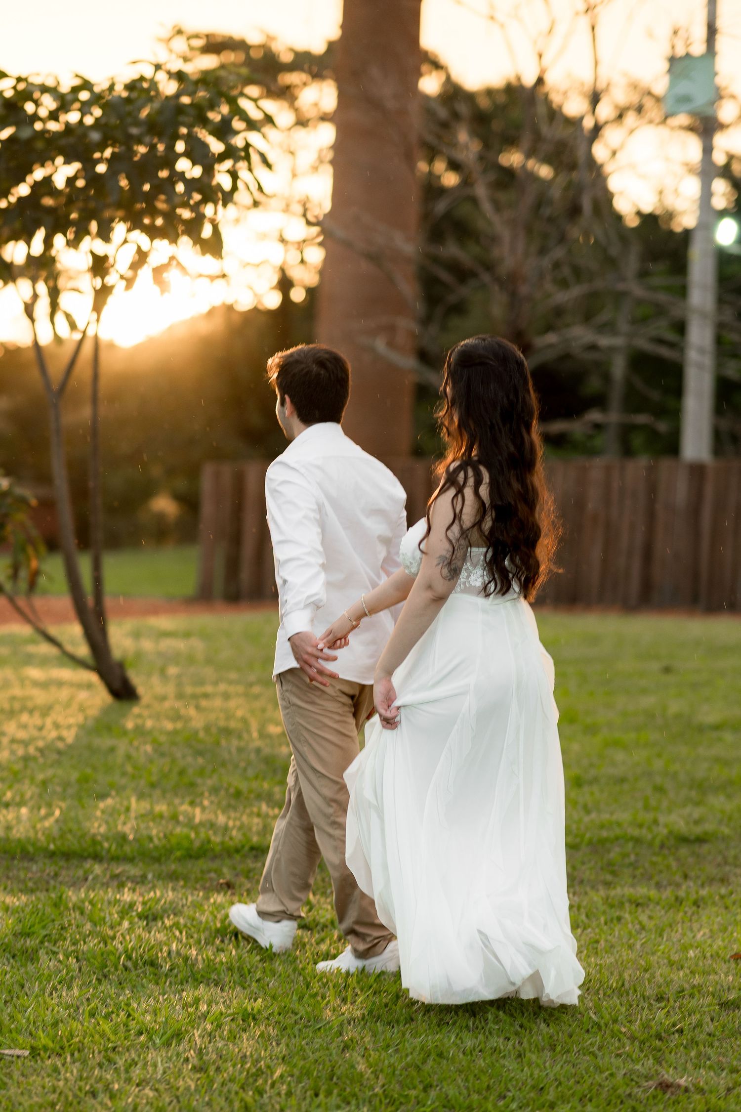 Couple in wedding attire holding hands walking through a sunlit garden at golden hour sunset.