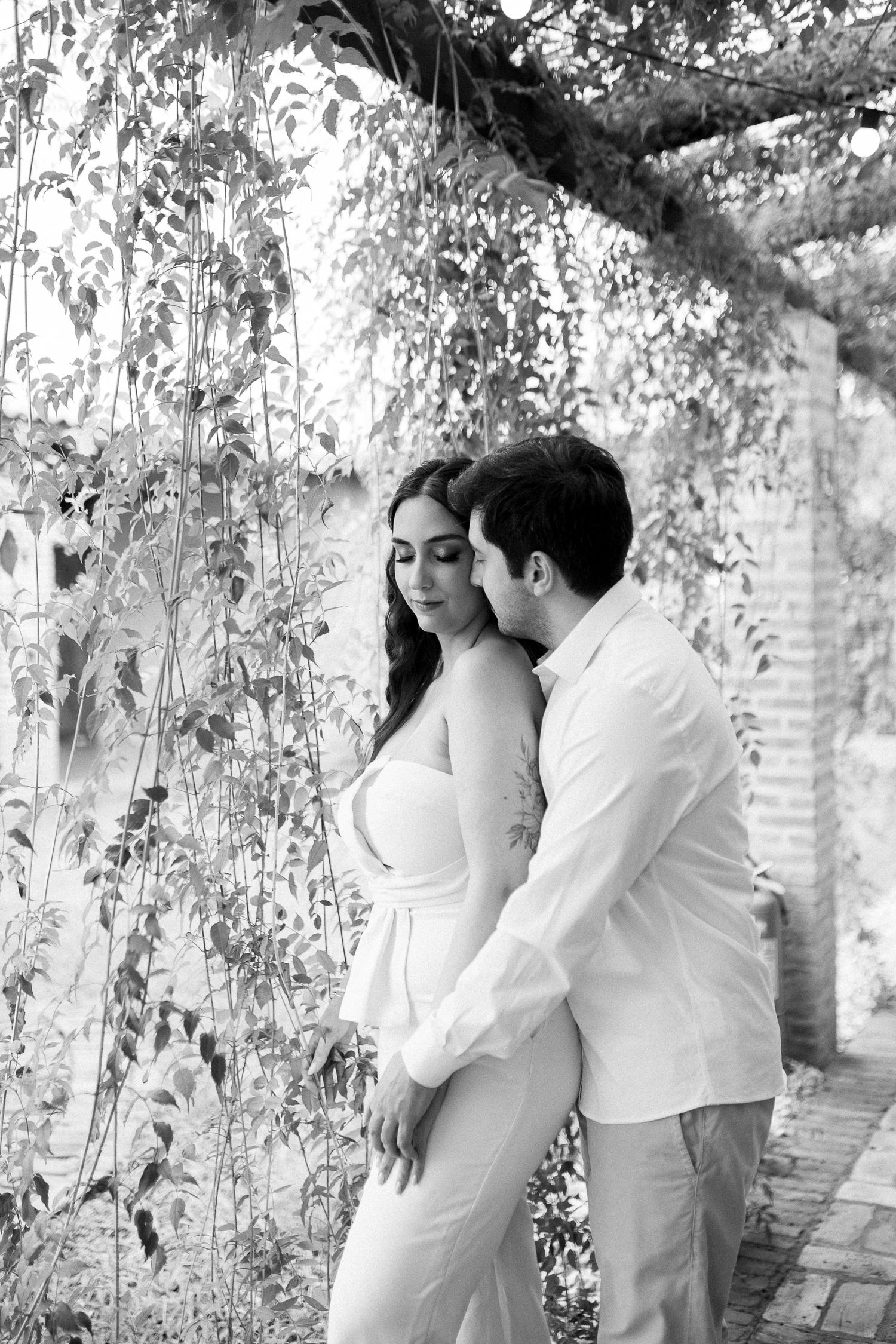 Black and white photo of romantic couple embracing under leafy pergola, woman in white dress, man in white shirt.