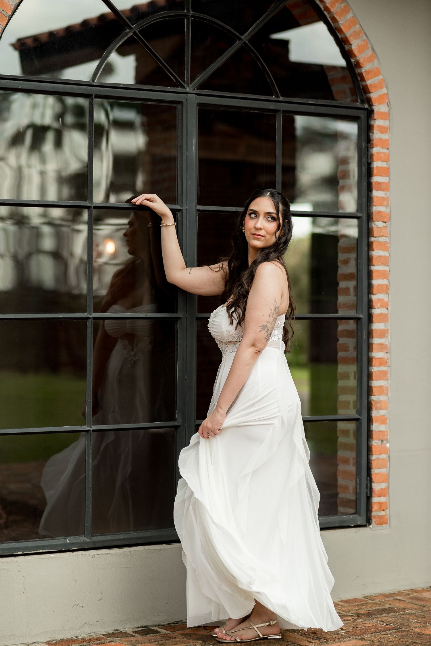 Bride in flowing white gown poses elegantly by arched window with brick frame.