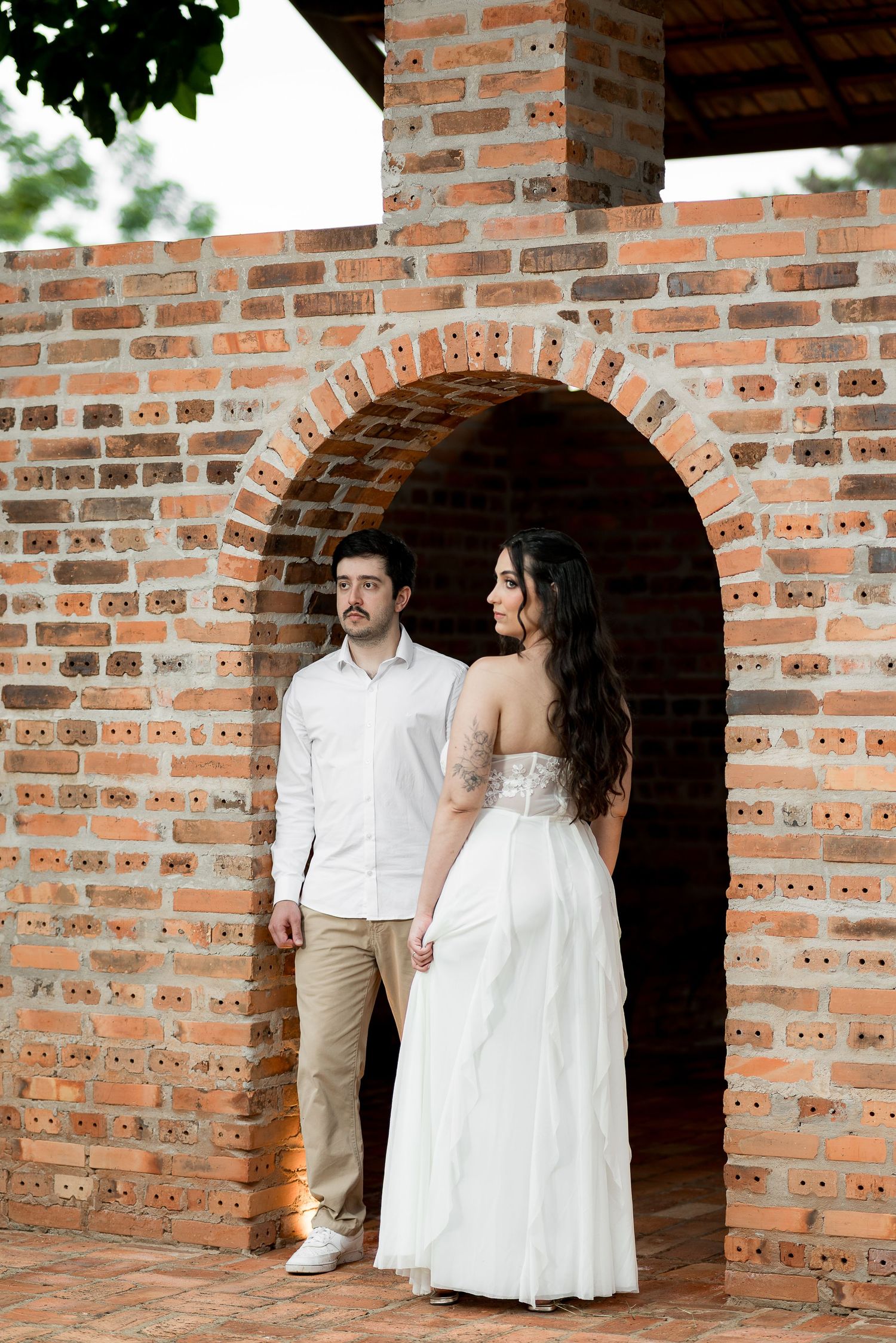 Bride in white gown and groom in white shirt pose under a rustic brick archway during an outdoor wedding photo session.