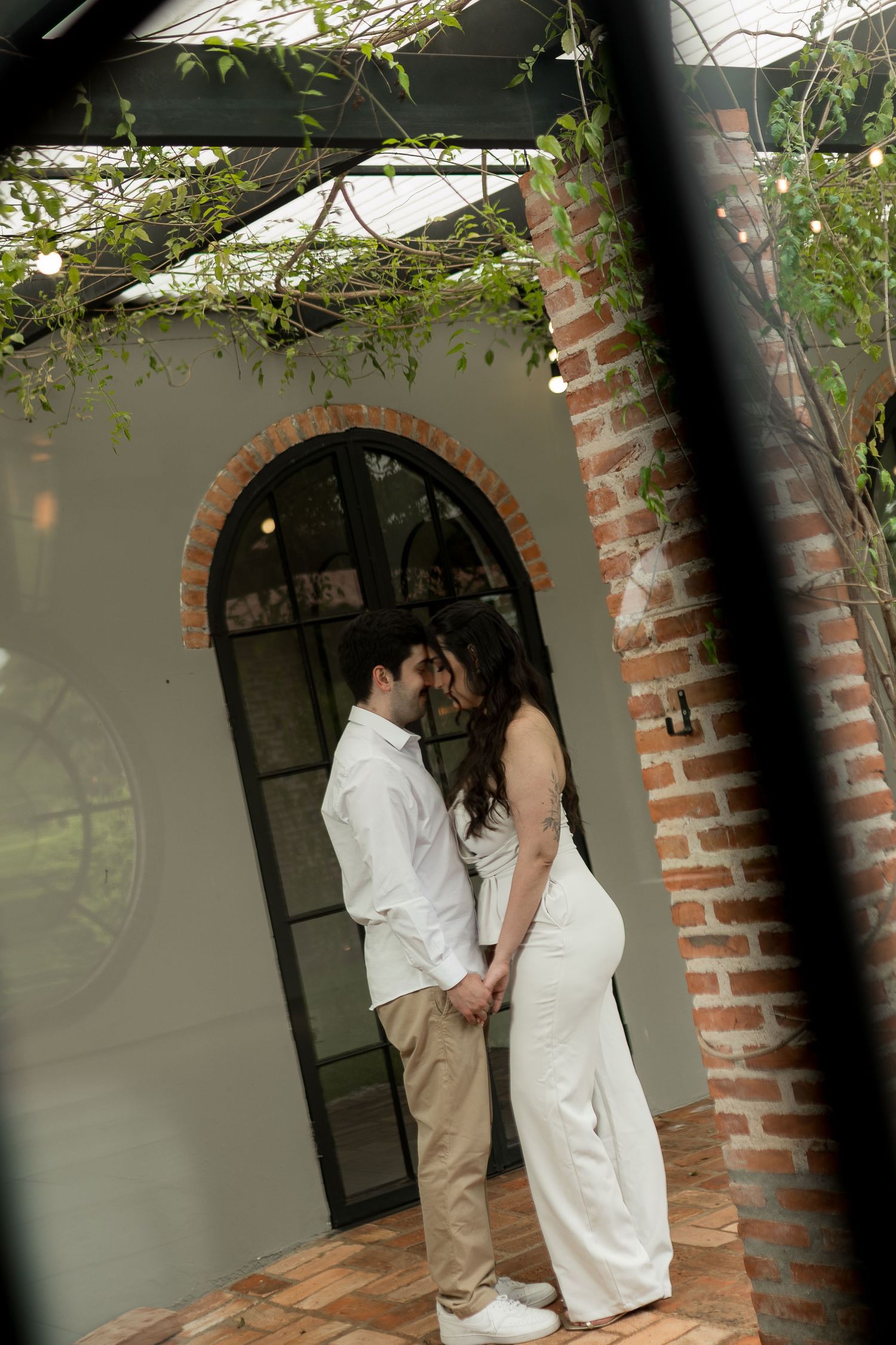 Couple in white outfits holding hands and sharing an intimate moment by a brick pillar and arched window.