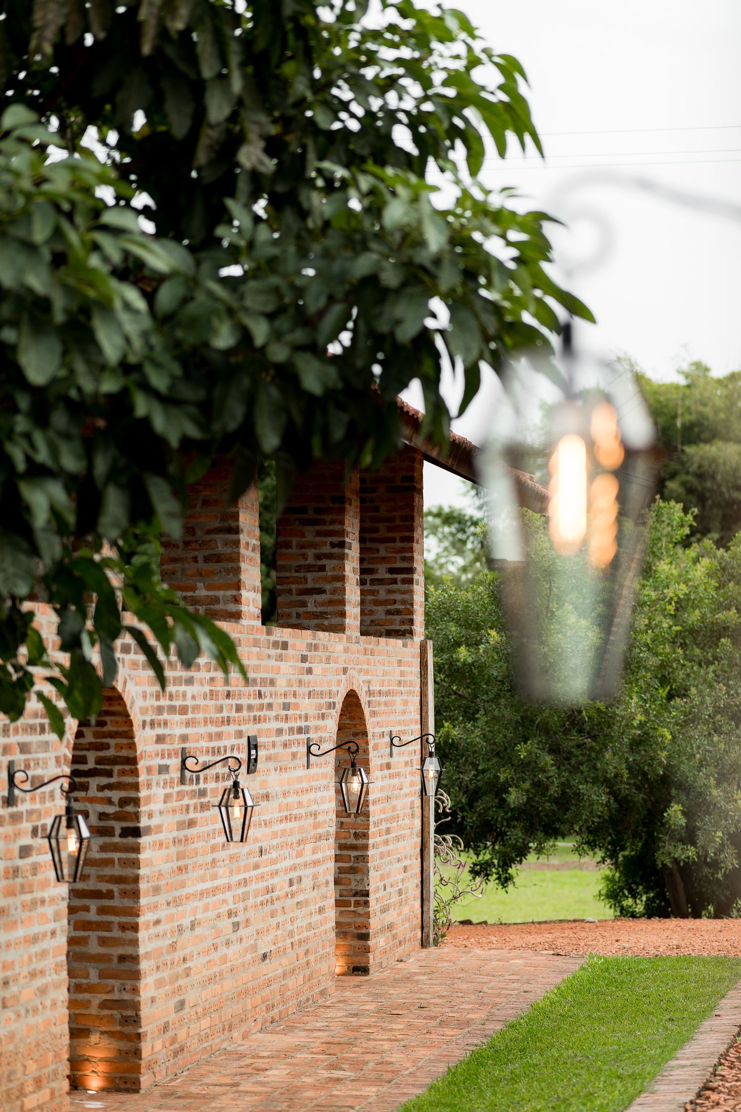Rustic brick building with arched alcoves, wrought iron lanterns, and lush green trees along a brick pathway.