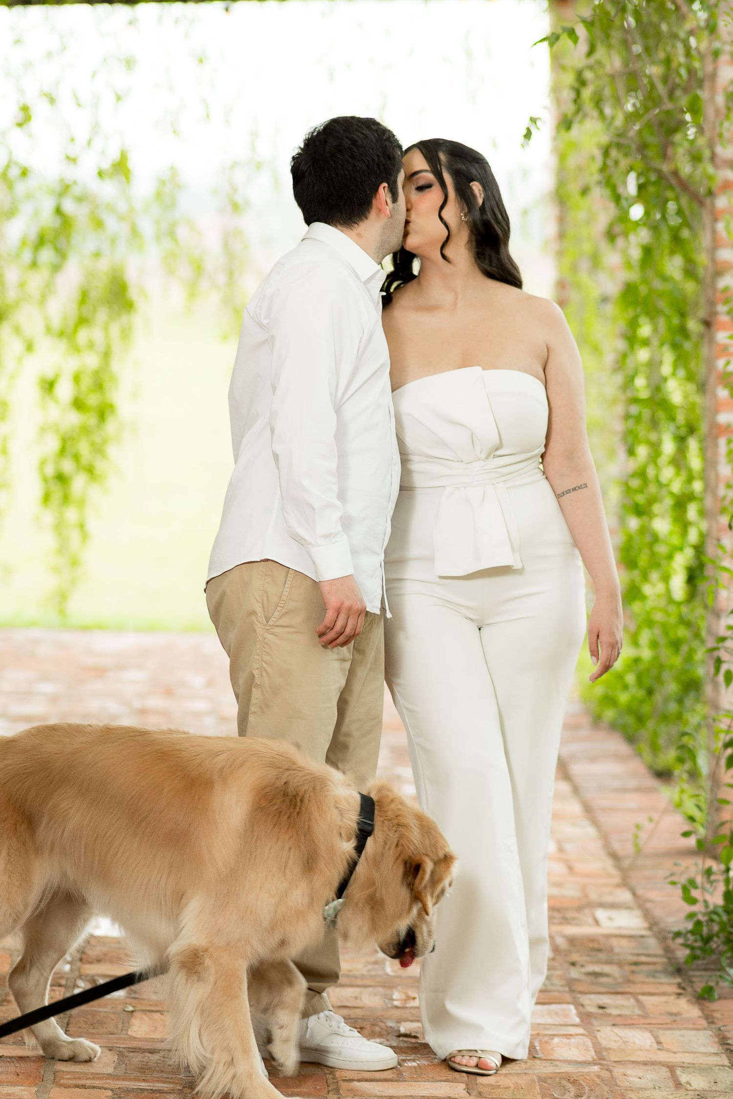 Couple sharing a kiss while walking their golden retriever on a brick path surrounded by lush greenery.