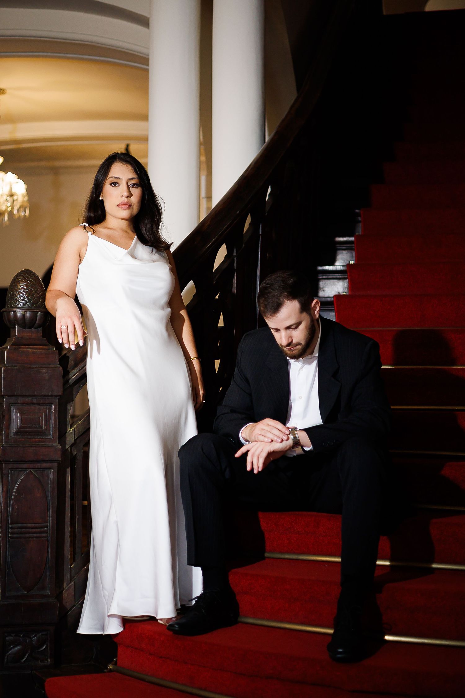 Elegant couple on red-carpeted stairs, woman in white gown standing, man in black suit seated examining her hand.