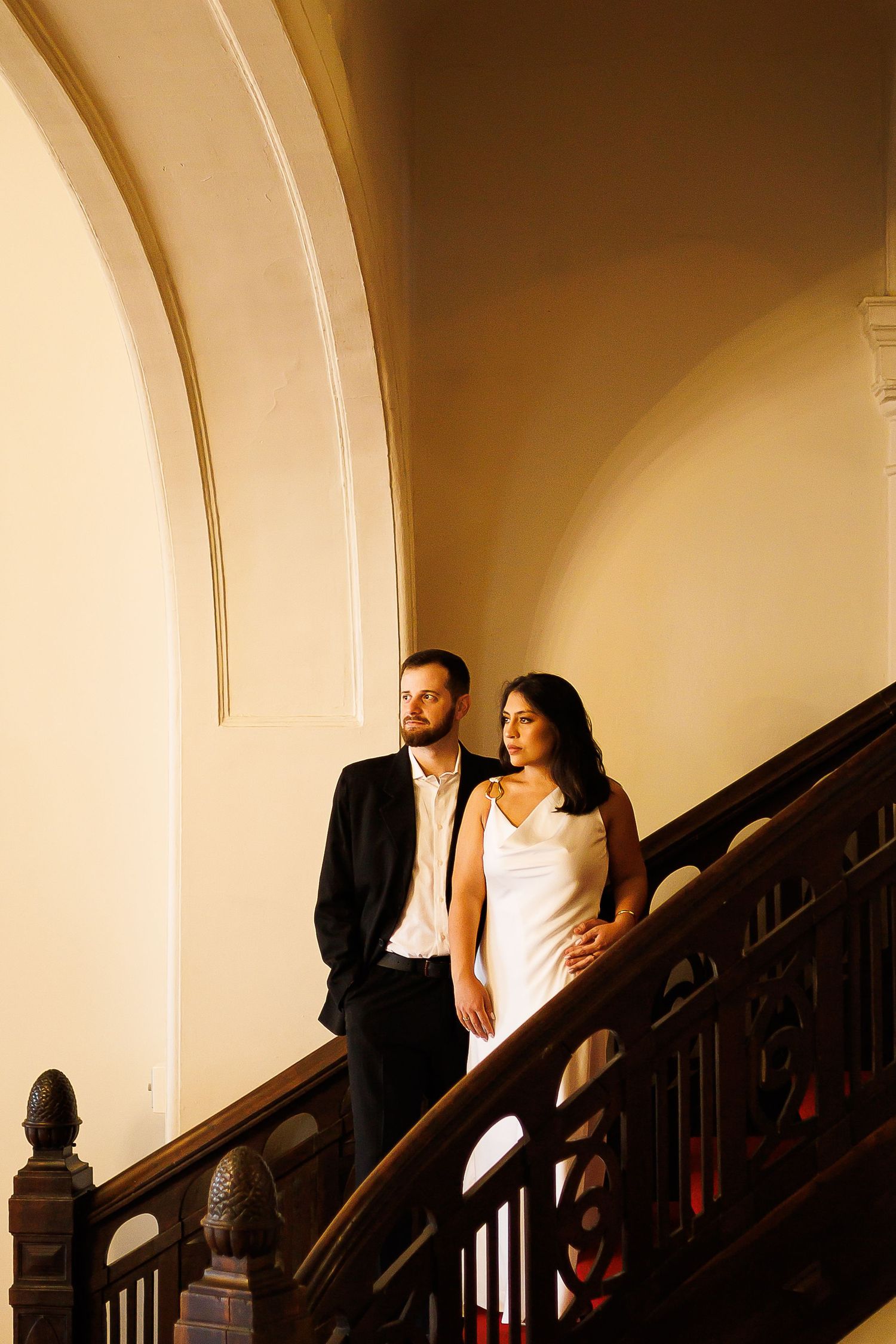 Elegant couple posing on ornate wooden staircase in grand building, man in suit, woman in white dress.