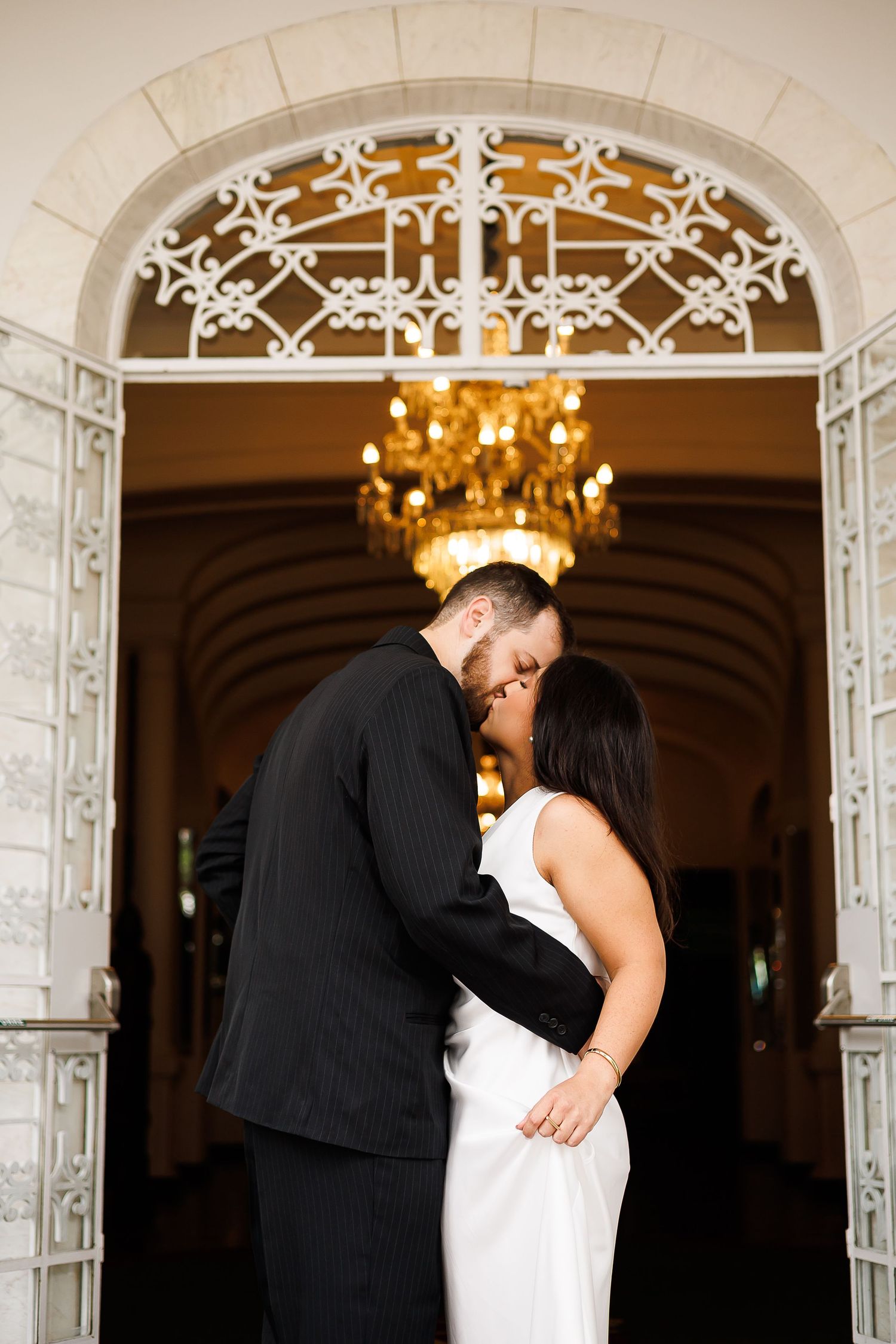 Couple sharing a romantic kiss in white dress and black suit beneath ornate arch with glowing chandelier.
