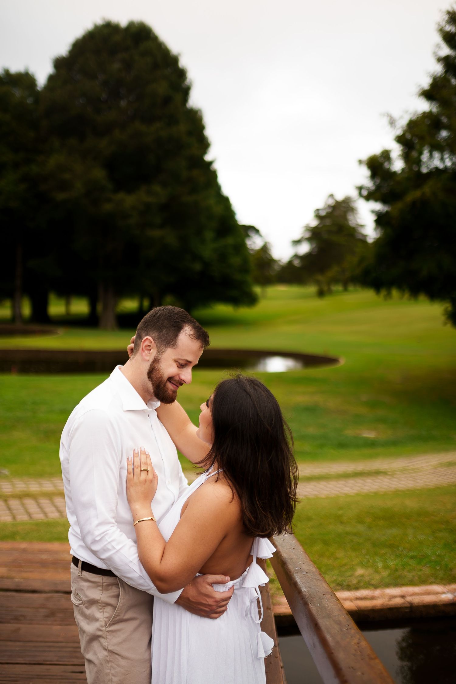 Couple in white embracing on a wooden bridge surrounded by lush green park trees and a pond.