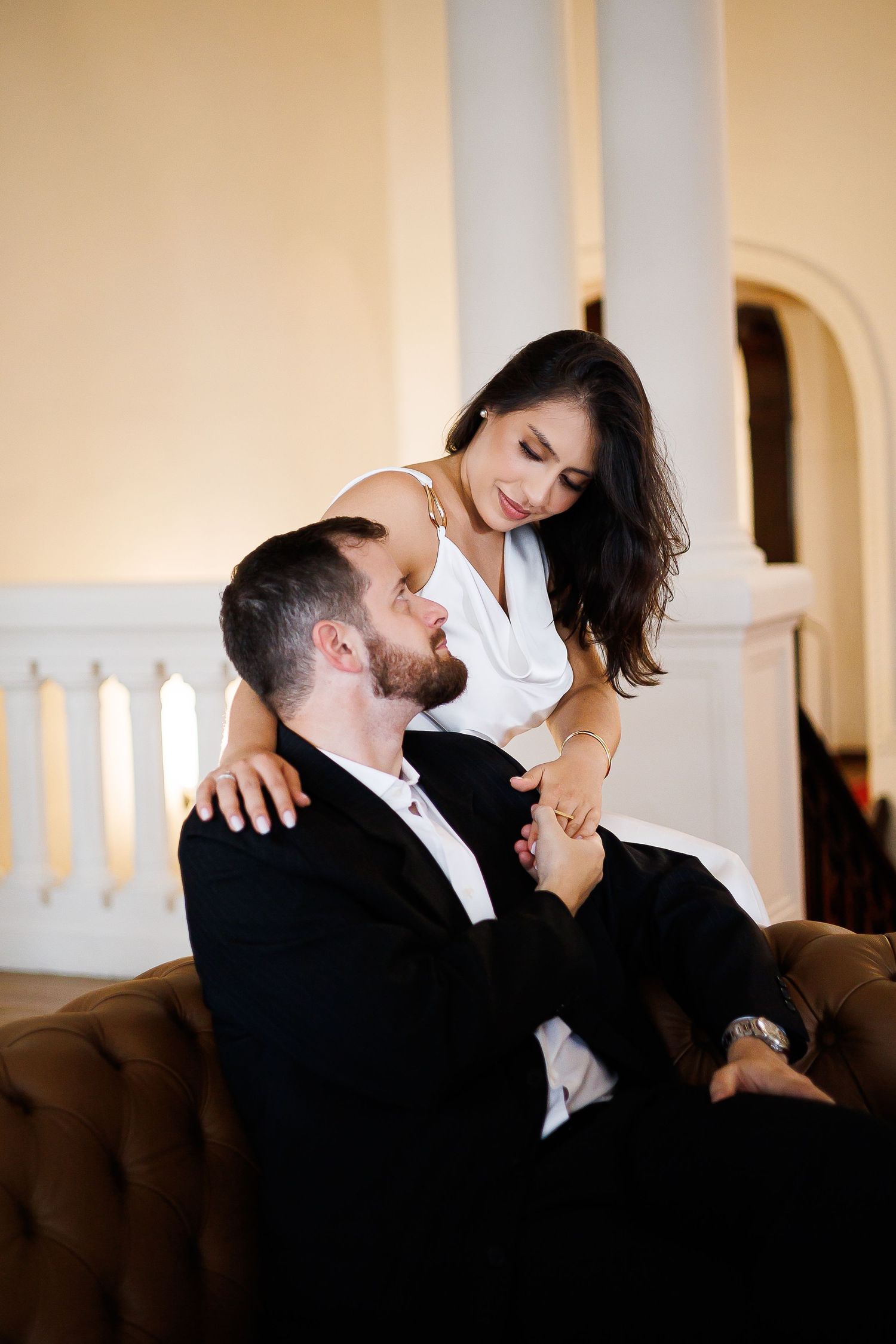 A couple shares a romantic moment, man in black suit seated on brown leather sofa, woman in white dress leaning over him.
