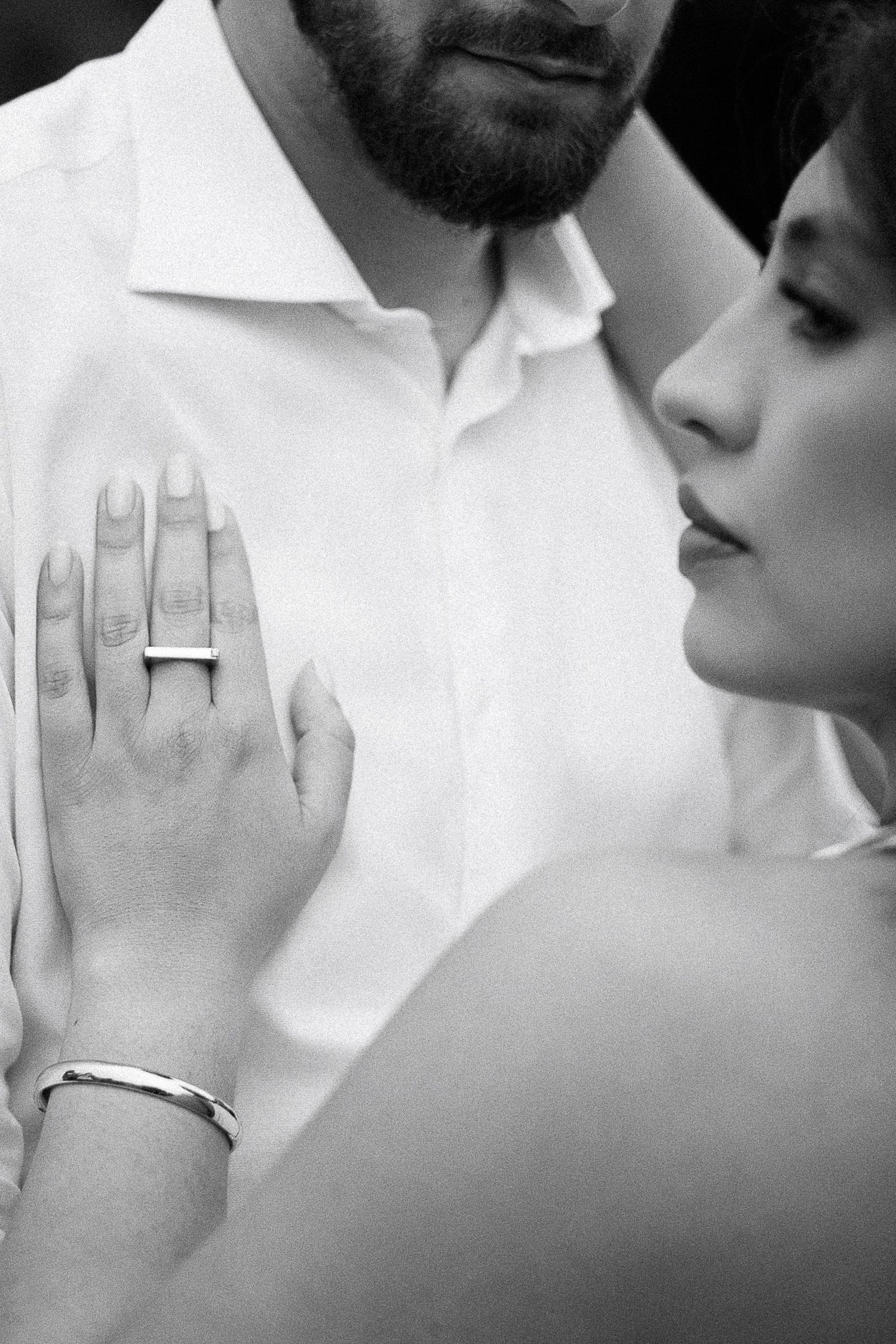 Black and white photo of a couple, woman showing engagement ring and bracelet while resting hand on mans white shirt.