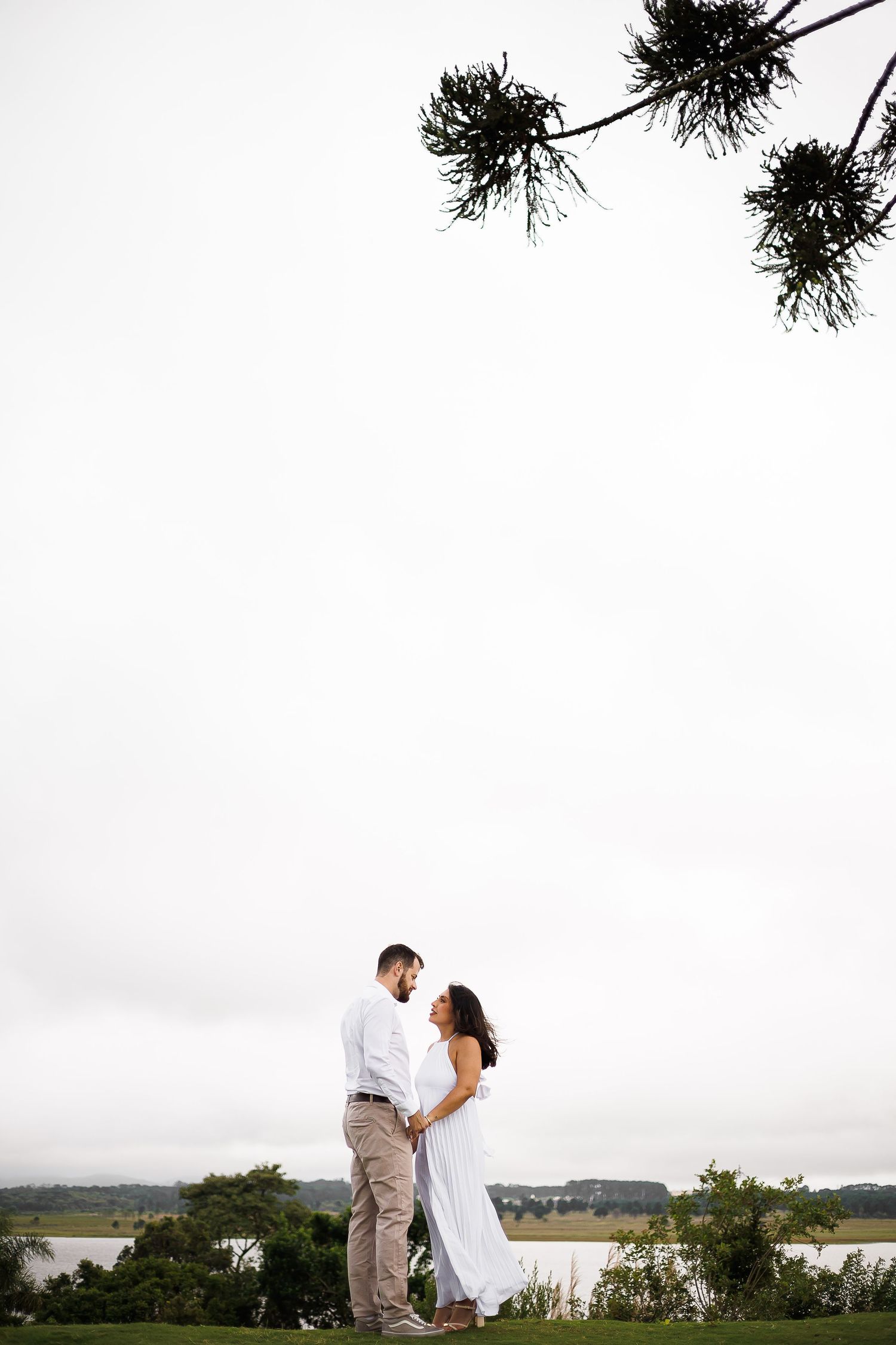 Couple in white attire standing face to face on green lawn with water view under pine branches on overcast day.