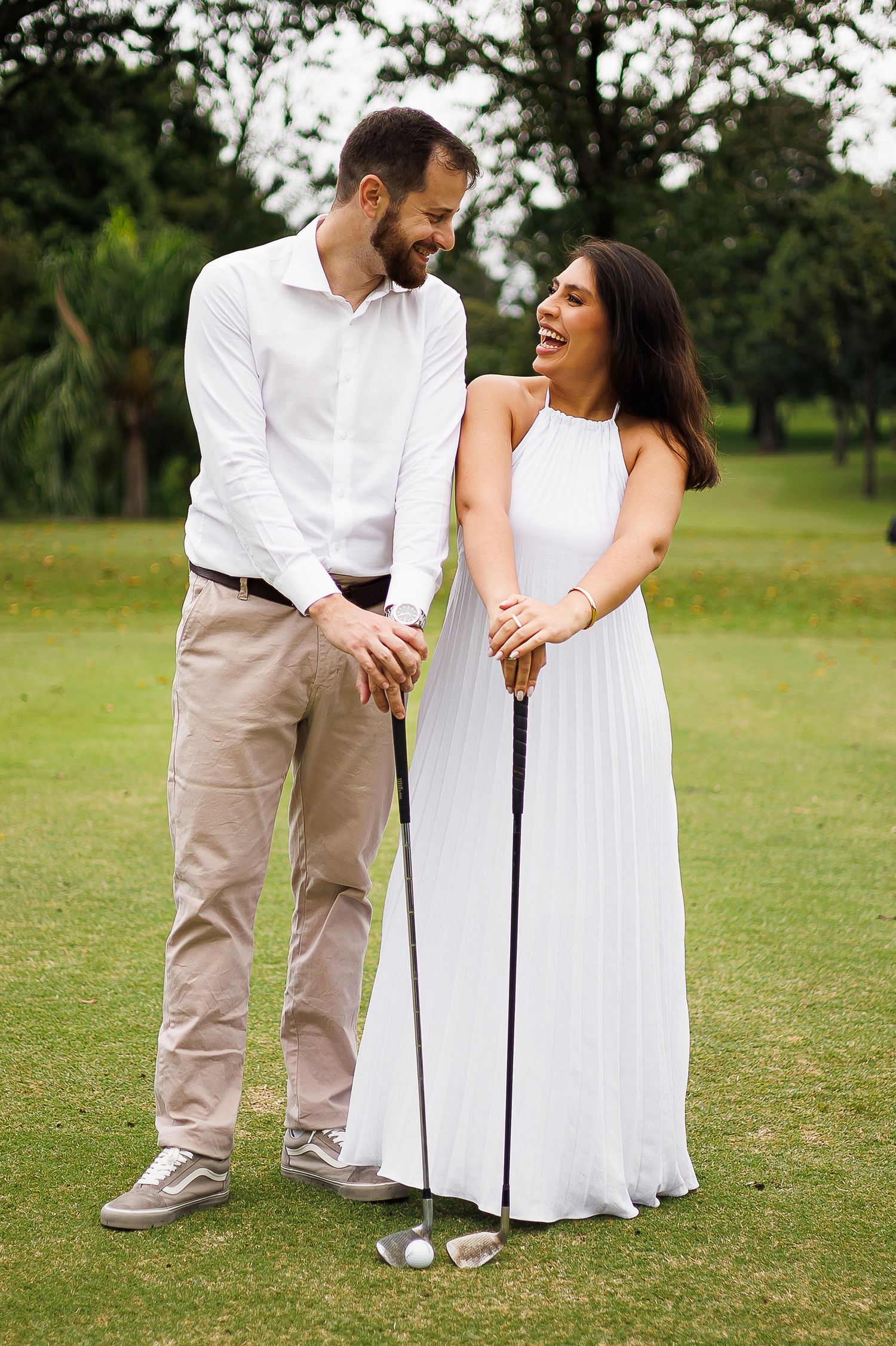 Couple laughing together on a golf course, woman in white dress holding a golf club, man in white shirt and khaki pants.