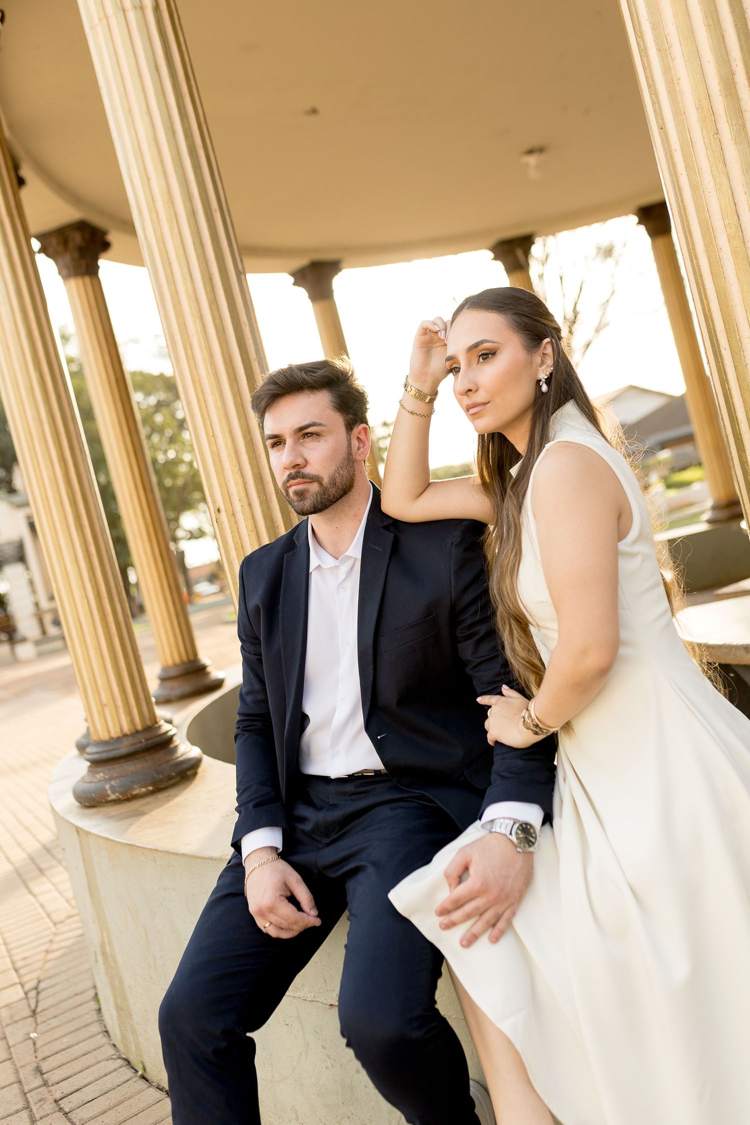 Elegant couple in formal wear posing at a classical columned pavilion, man in black suit and woman in white dress.