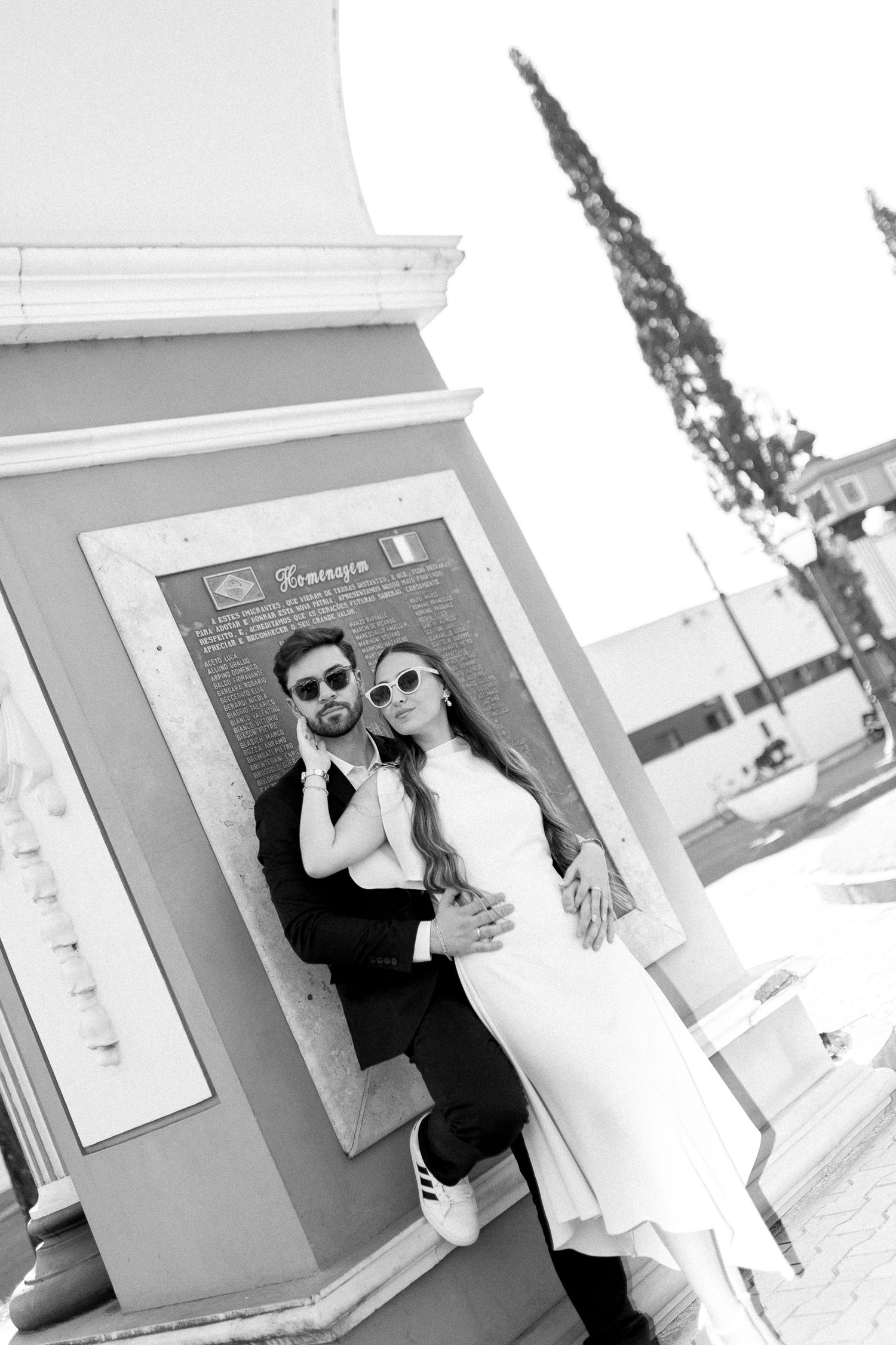 Black and white photo of a stylish couple in sunglasses embracing near a monument plaque outdoors.