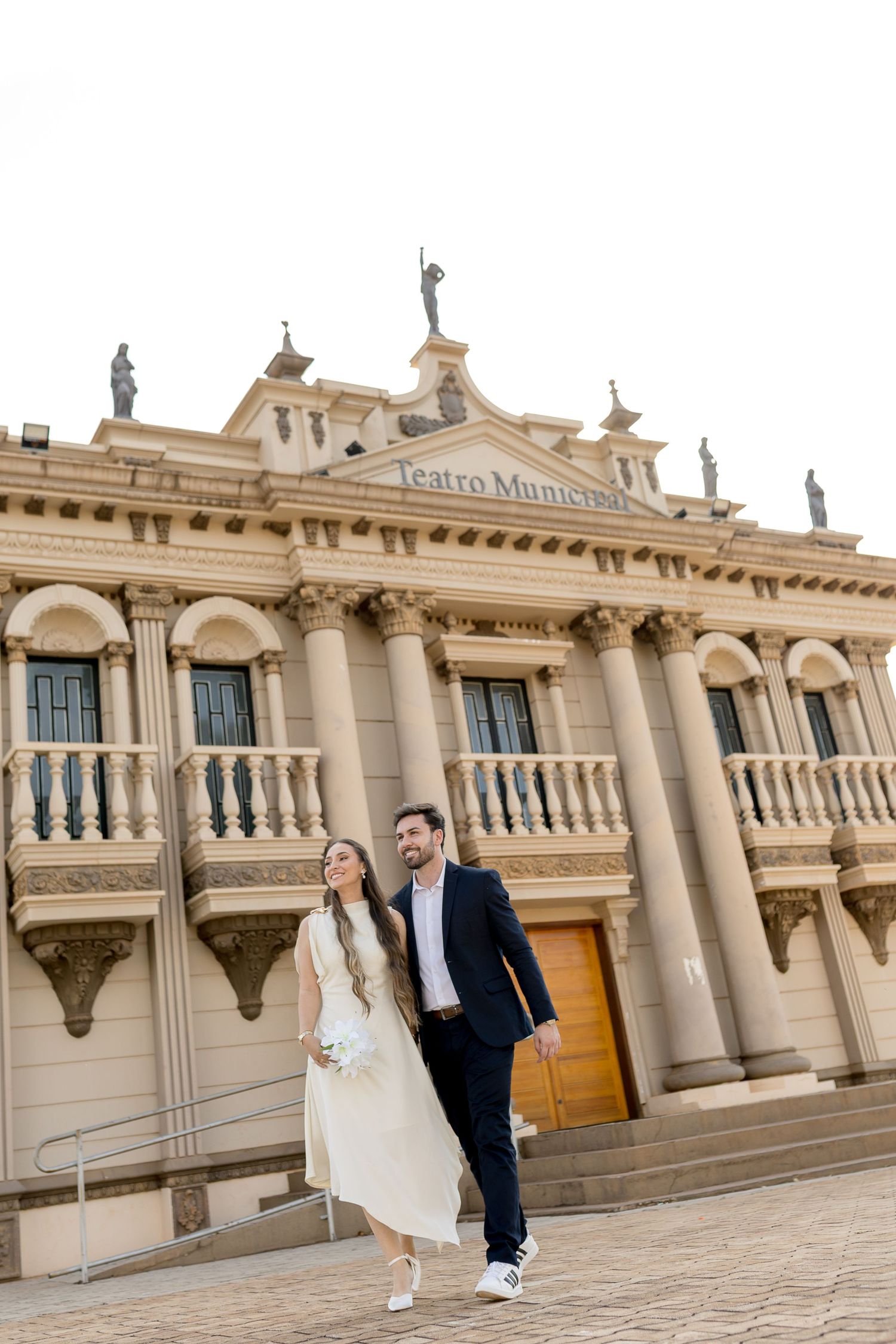 Couple in wedding attire walking in front of the historic Teatro Municipal building with classical columns.