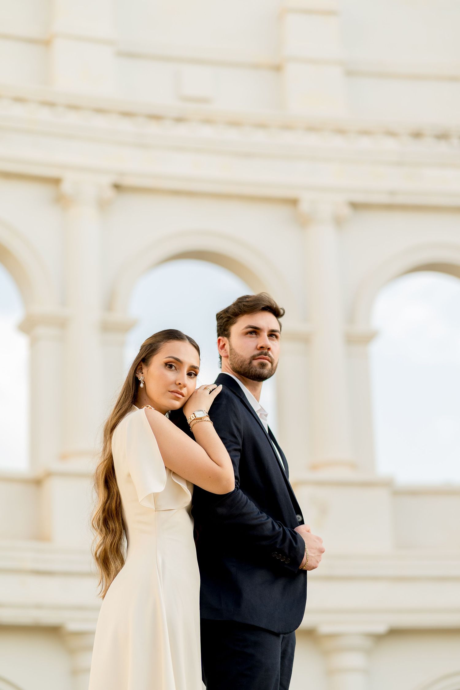 Elegant couple in formal attire posing in front of classical arched architecture, woman in white dress, man in black suit.