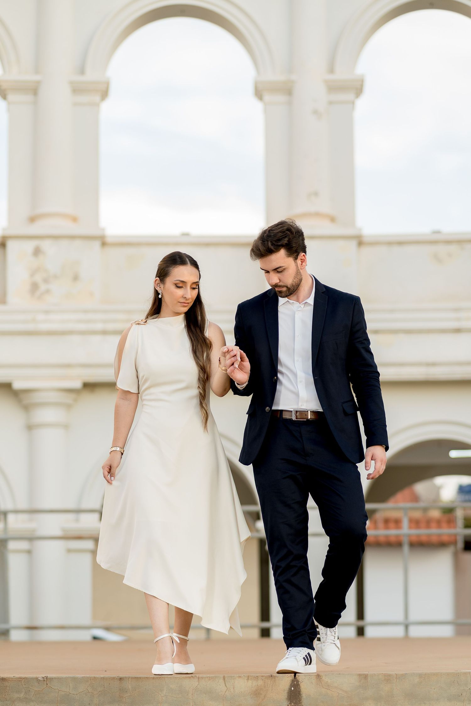 Elegant couple walking together, woman in white dress and man in navy suit, posing before grand arched building exterior.