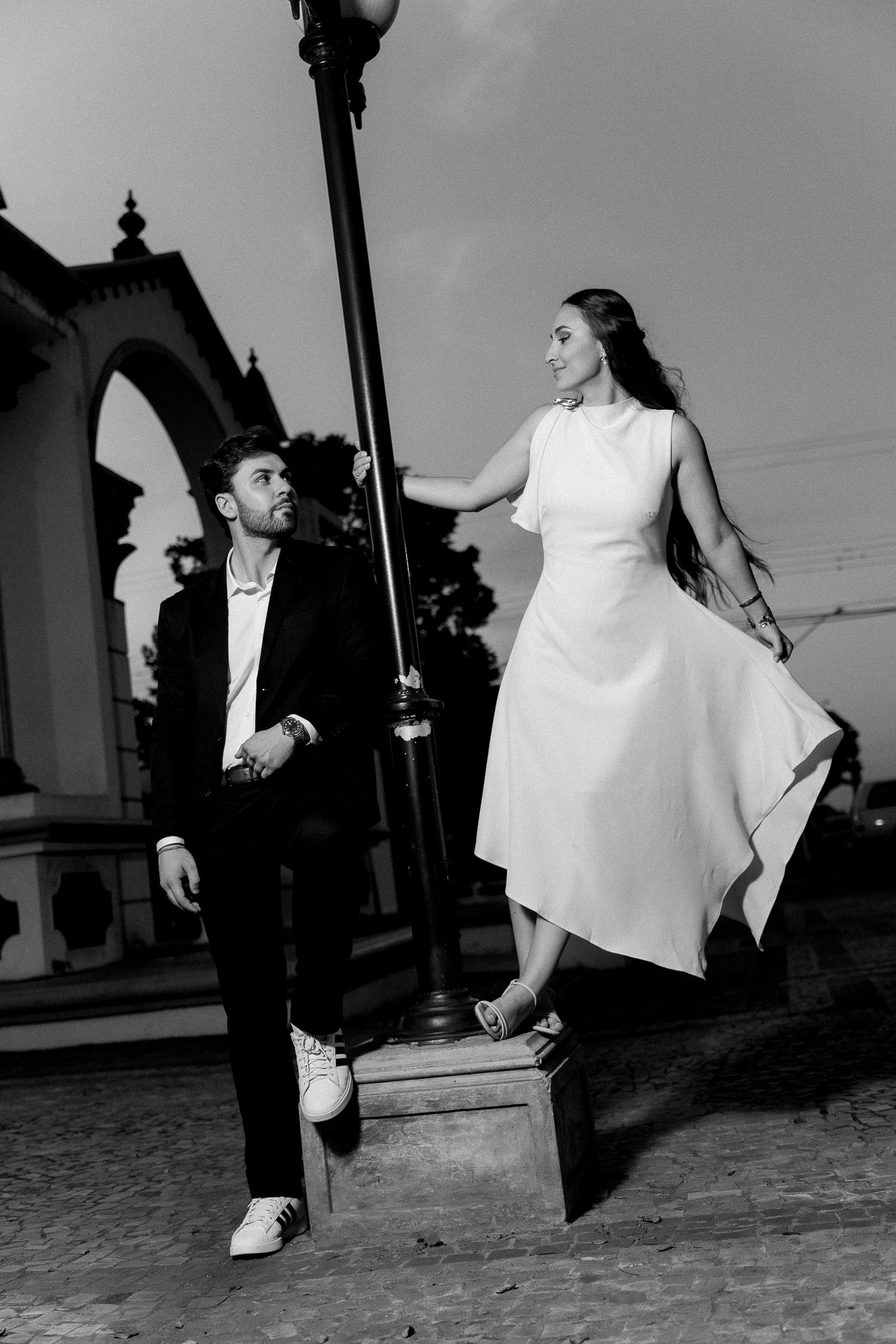 Black and white photo of elegant couple near street lamp, woman in white dress standing on pedestal, man in suit looking up.
