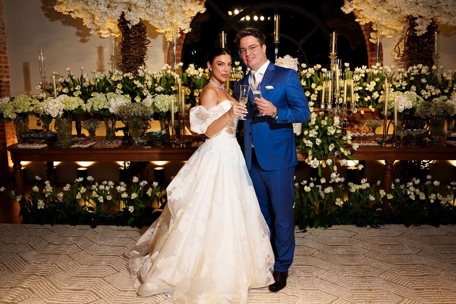 Bride and groom toast with champagne at elegant floral wedding reception with white blooms and candlelit backdrop.