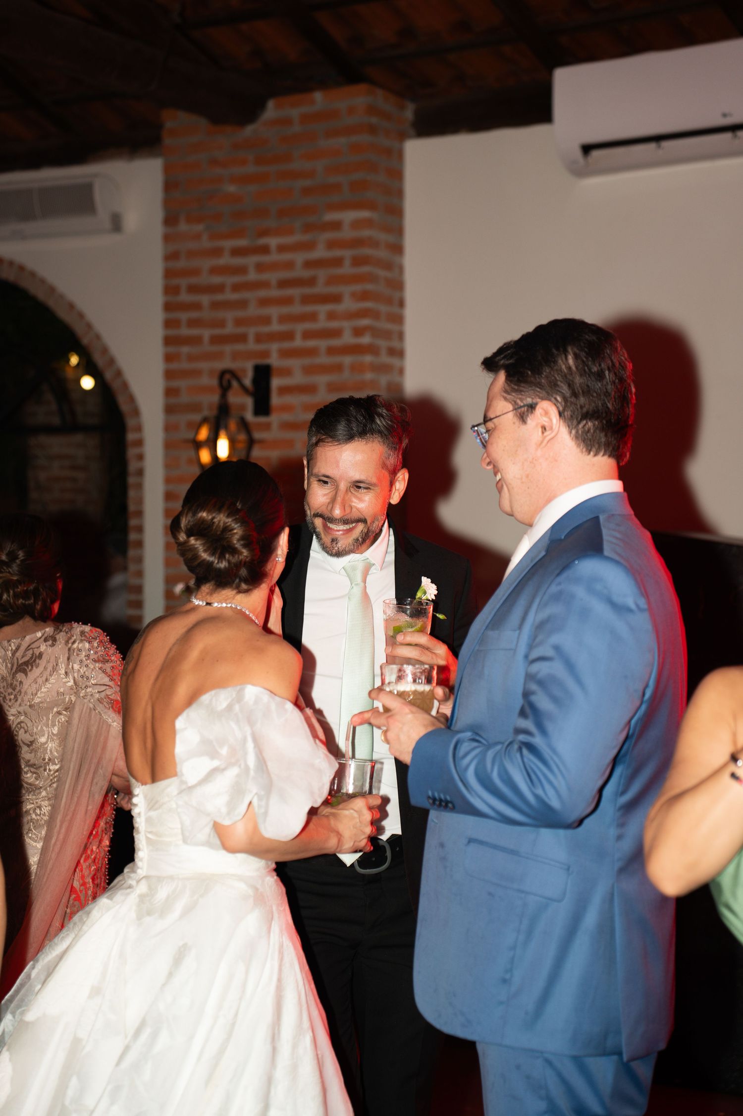 Bride and groom in wedding attire clinking champagne glasses with a guest in a blue suit at an elegant indoor reception.